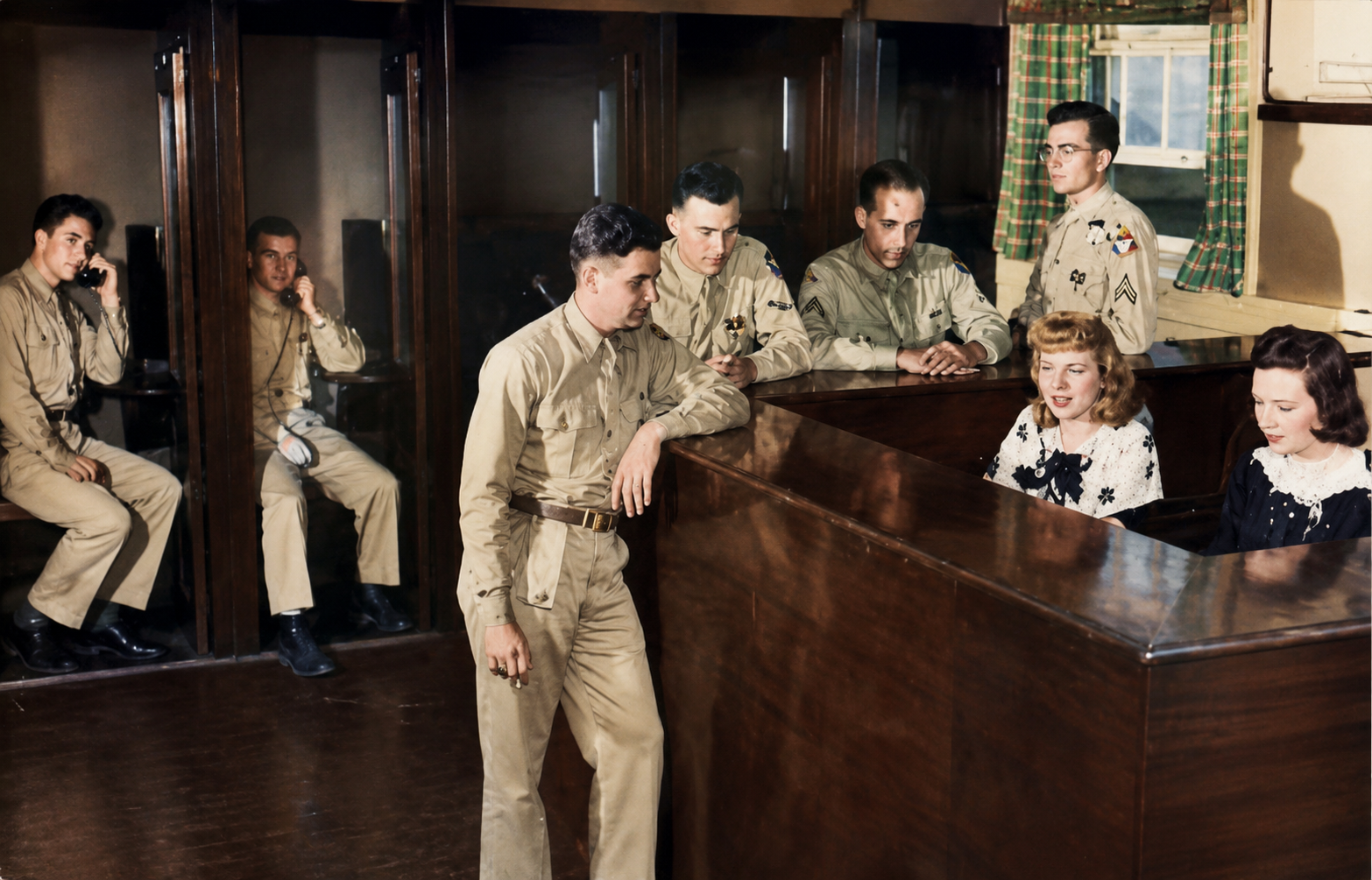 A courtroom scene with soldiers in uniform, two young women sitting at a bench, and a soldier standing in front of them. The soldiers are engaged with phones and attentive, while the women are looking towards the soldier.