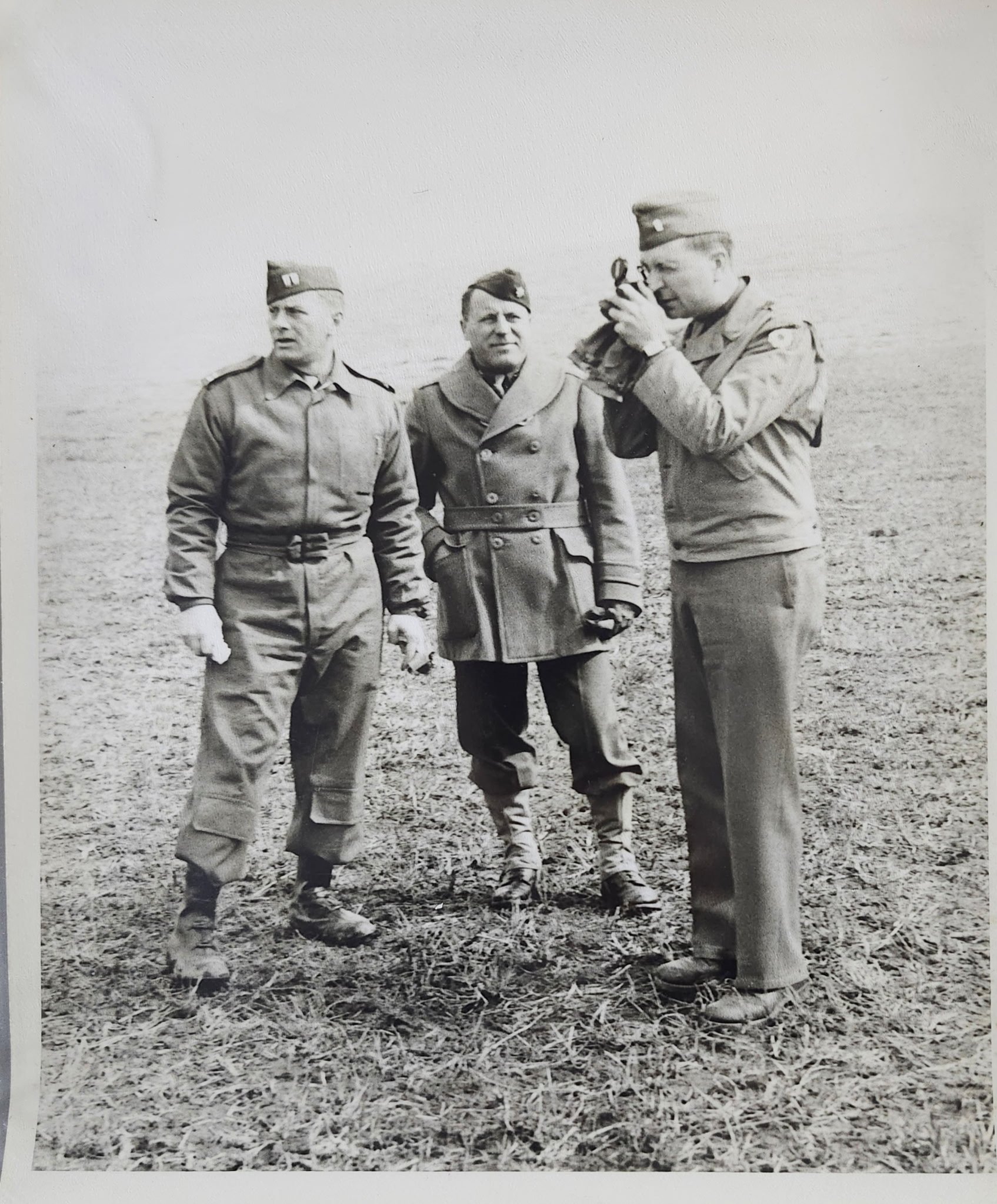 Three soldiers in uniform standing outdoors on a field, with one of them aiming a pistol and the others standing nearby.