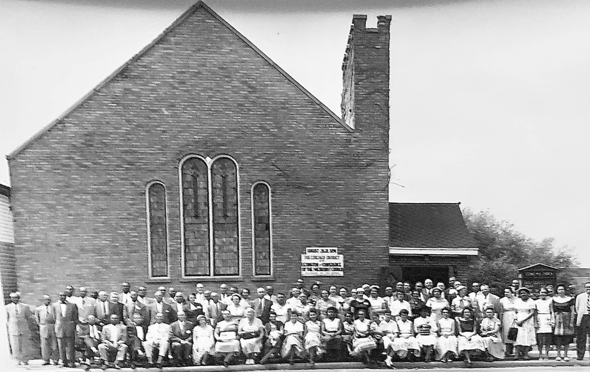 Black and white photo of a large group of people standing in front of a brick church building with tall windows and a steeple, possibly from the early to mid 20th century.