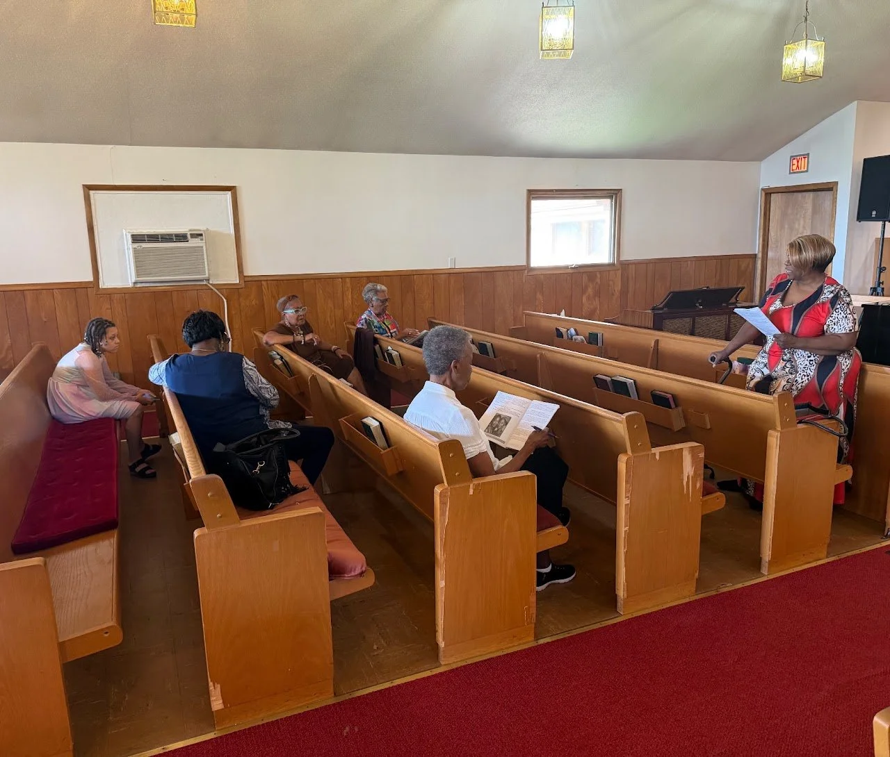 A woman in a red patterned dress leading a small group in a church or community room, with six people seated on wooden pews listening to her, some holding papers or books.