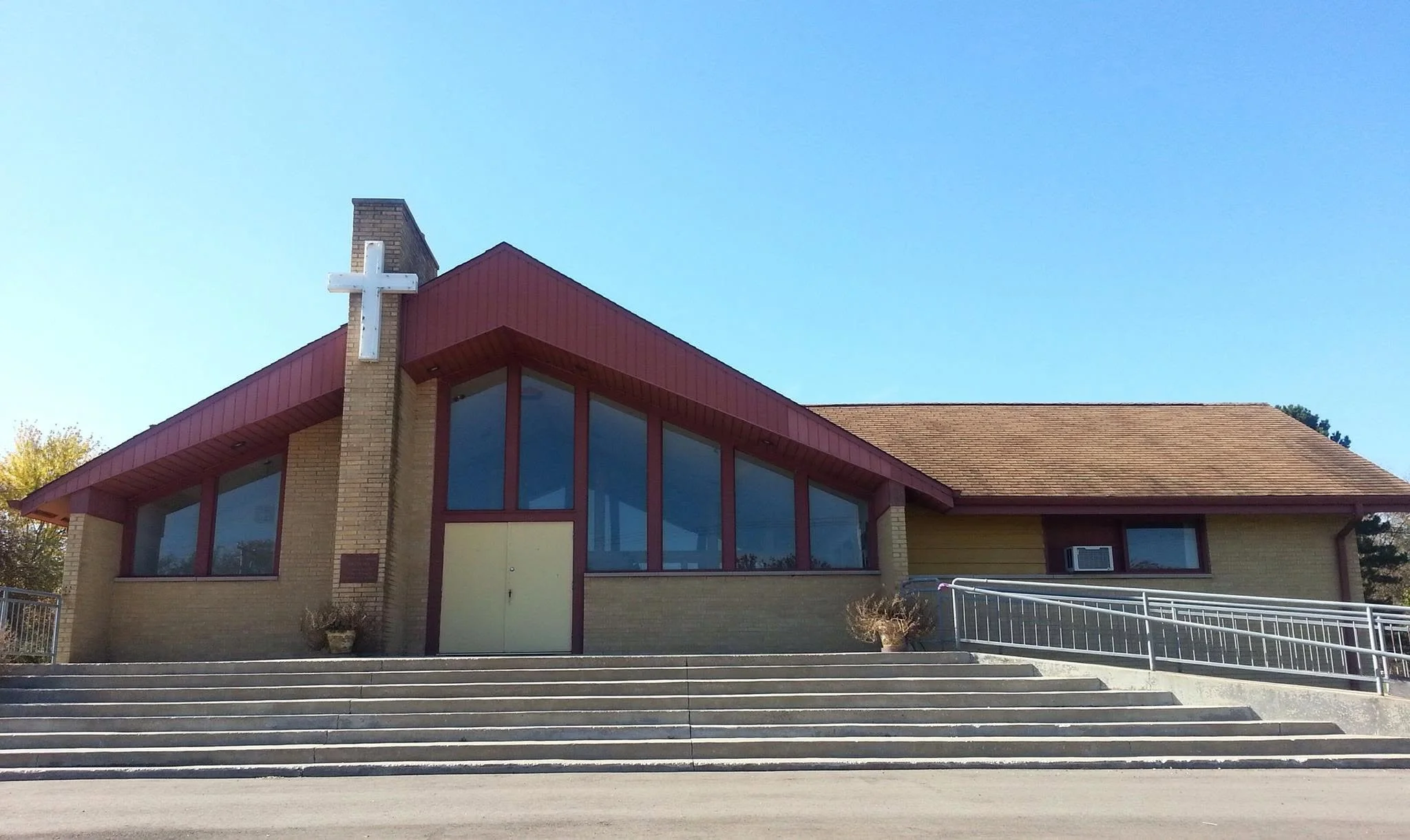 A church building with a cross on top, large windows, brick and yellow exterior, and a sloped roof under a clear blue sky.