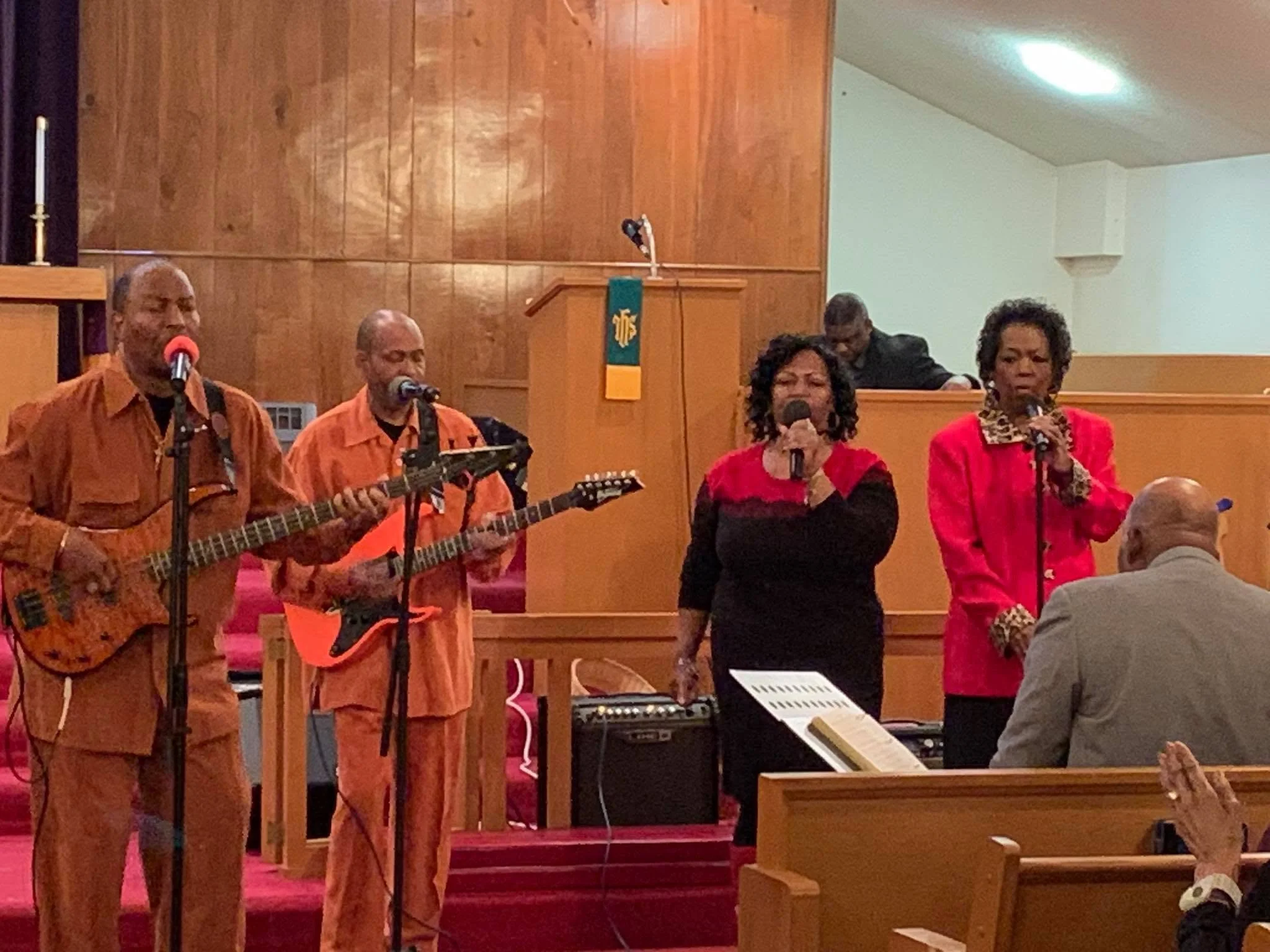Four Black gospel singers, three women and one man, singing into microphones on a stage with a wooden backdrop. The women are dressed in red and patterned clothing, and the man is dressed in an orange-brown shirt and pants. There's a person playing a guitar and a keyboard visible, with additional people in the background.