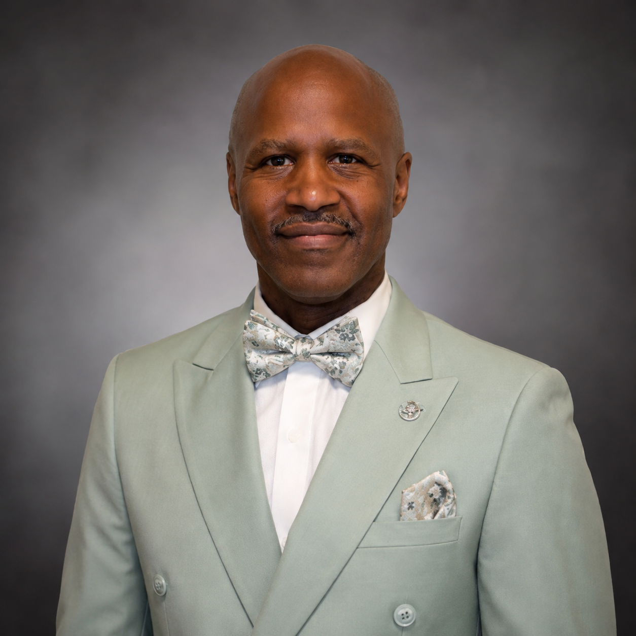 A portrait of an older man with dark skin, bald head, wearing a light green suit, patterned bow tie, and a matching pocket square, smiling slightly against a gray background.