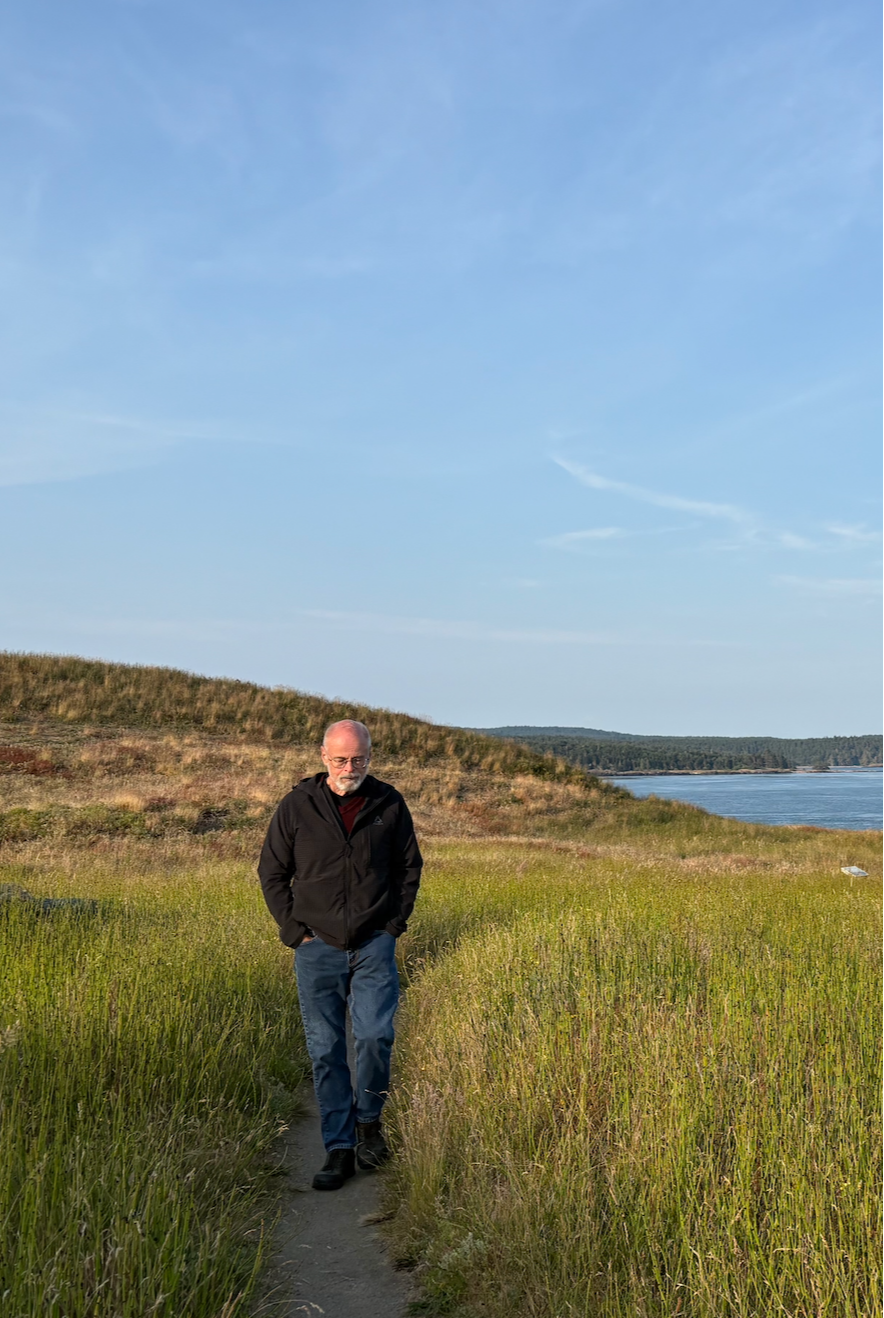 A man walking along a narrow dirt path through tall green grass with a lake and grassy hills in the background under a blue sky.