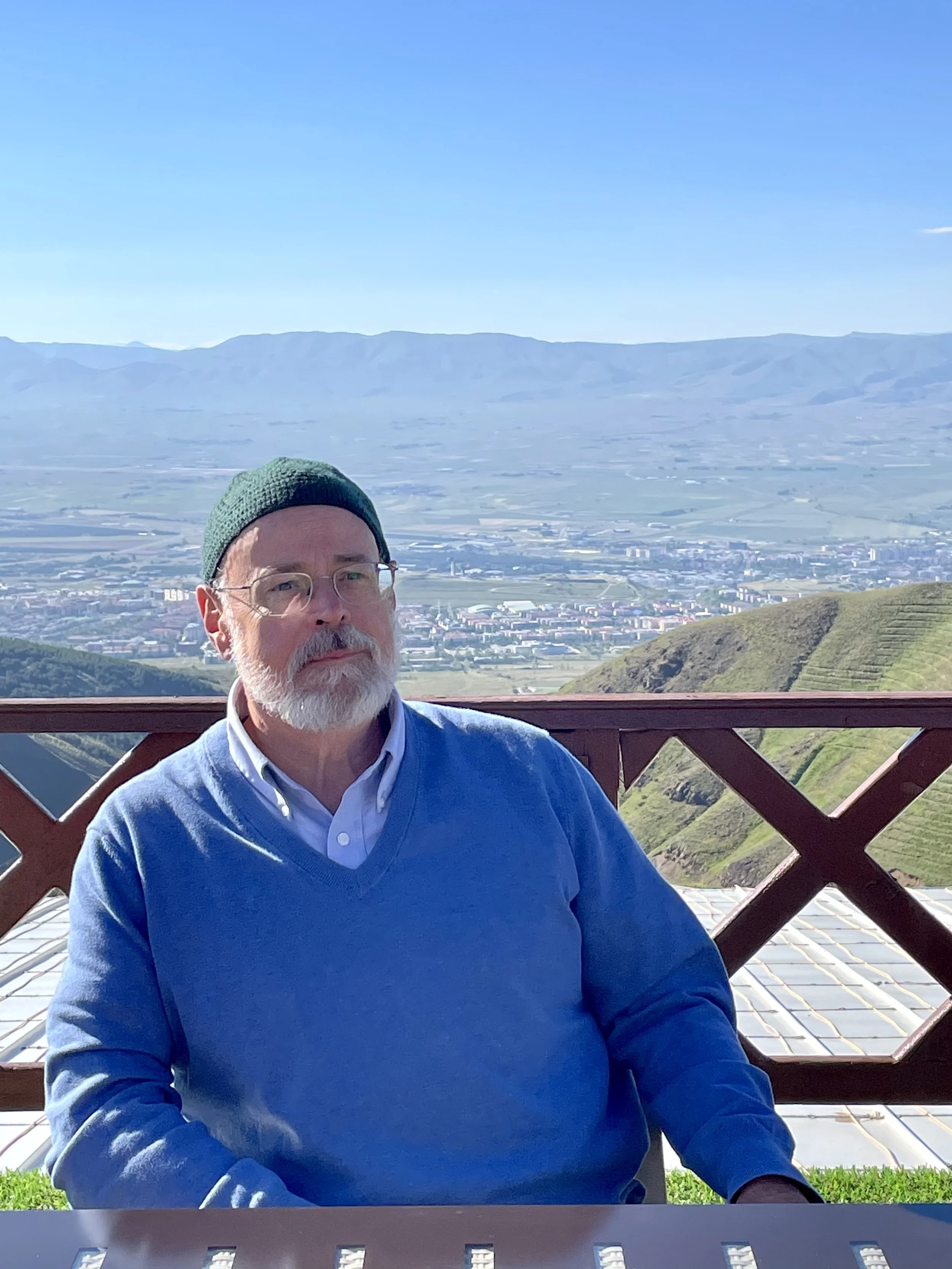 Timothy Burley with glasses, a gray beard, and a green beanie sitting outdoors on a wooden balcony with a scenic mountainous landscape and a town in the background.