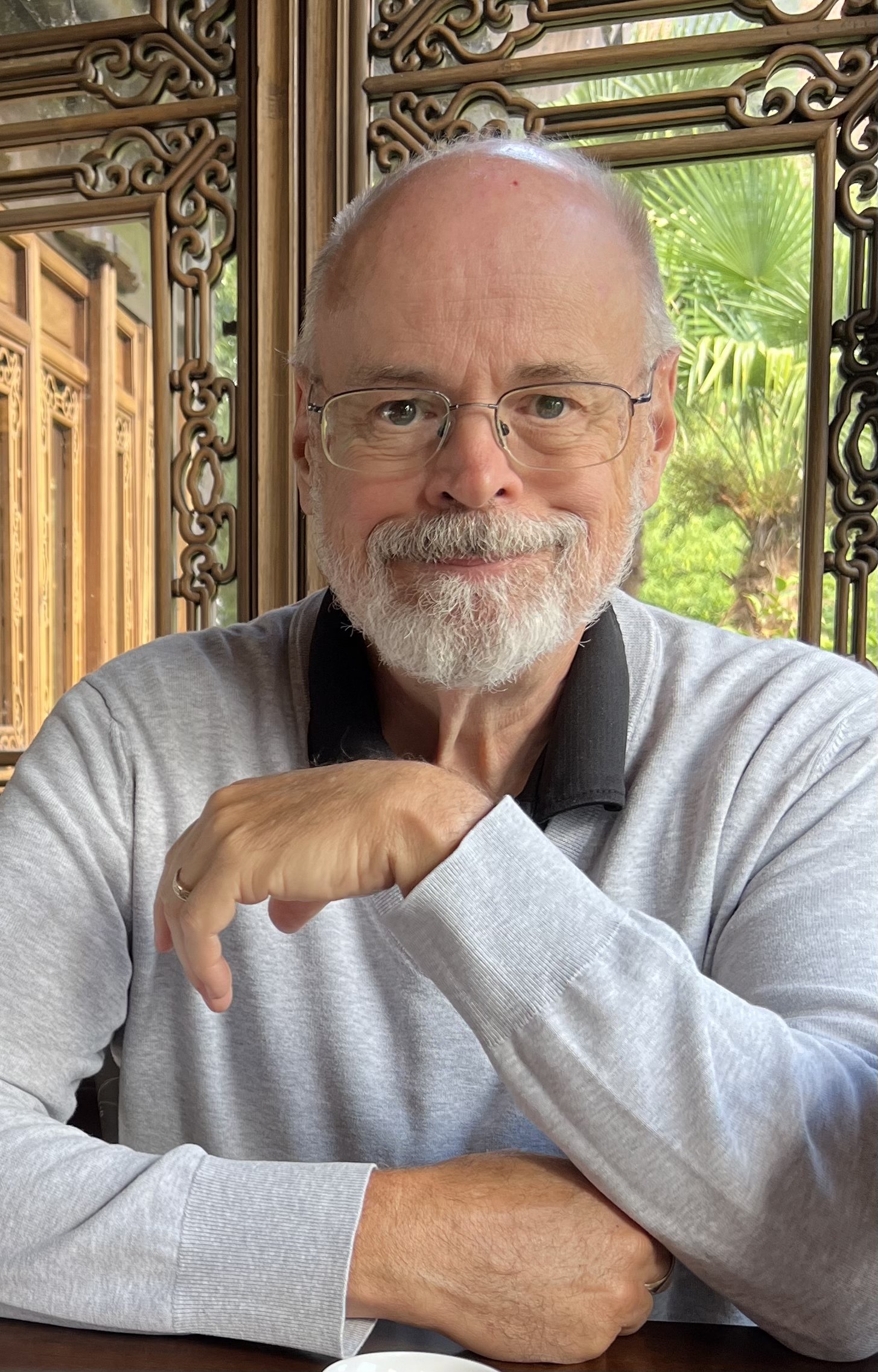 The author, Timothy Burley, with glasses, a gray beard, and a friendly smile, sitting at a table in a traditional wooden and bamboo setting with lush green plants outside.