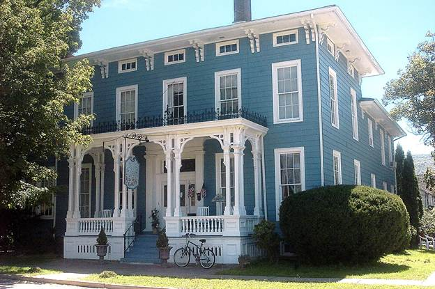A large, blue, two-story house with white trim and a wraparound porch with ornate columns, located in a residential neighborhood with trees and bushes.
