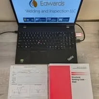 Desk with a laptop displaying Edwards Welding & Inspection LLC, alongside a WPS, and a AWS D1.1 code book.