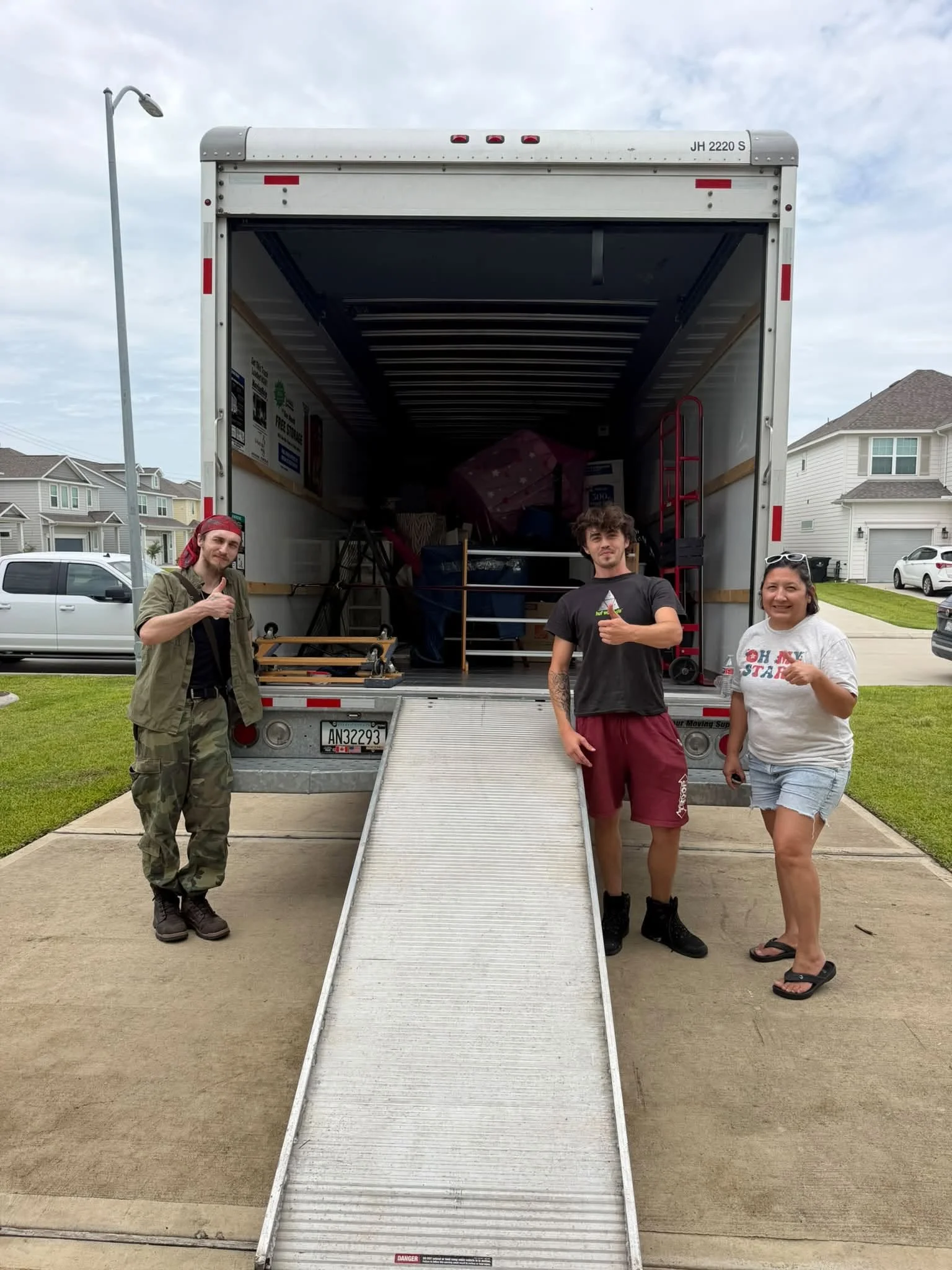 Three people standing in front of an open moving truck with a ramp, in a residential neighborhood, during daytime.