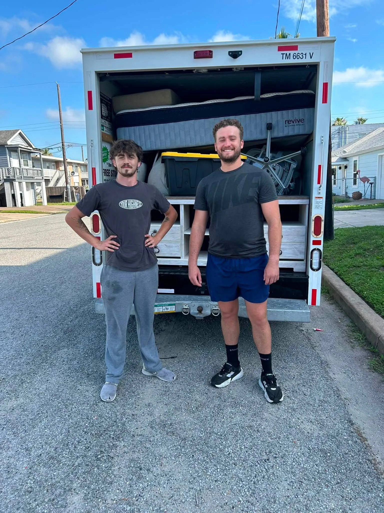 Two young men standing in front of a moving truck with packed belongings, smiling outdoors on a sunny day.