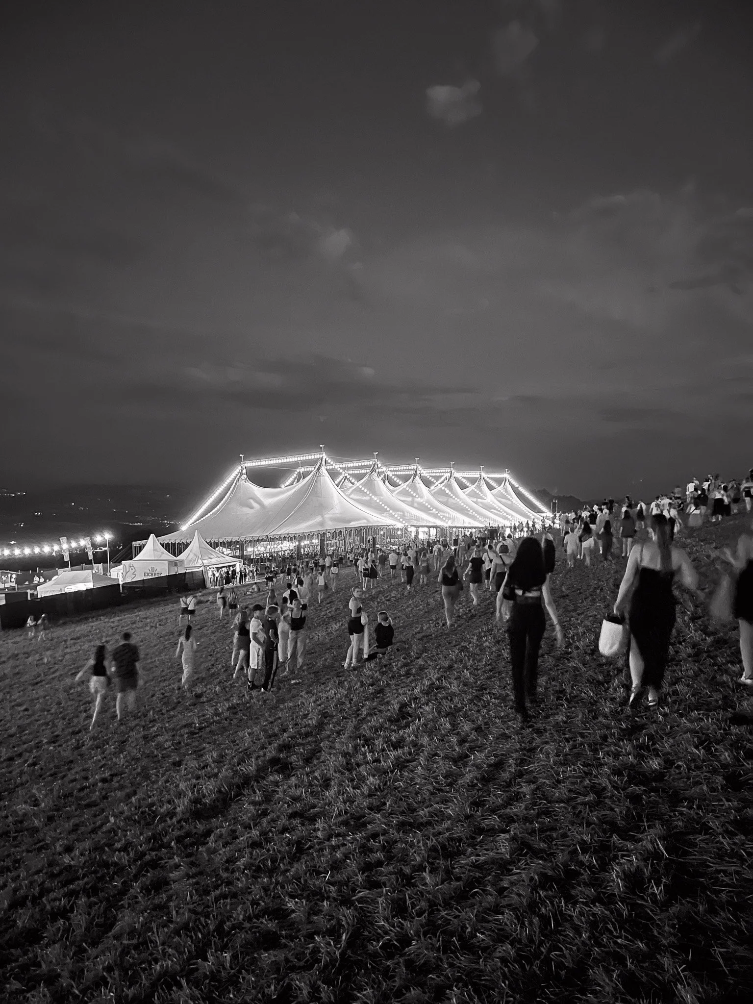 Das beleuchtete Zelt des Gurtenfestivals auf einer Wiese mit vielen Menschen, die umhergehen, bei Nacht.