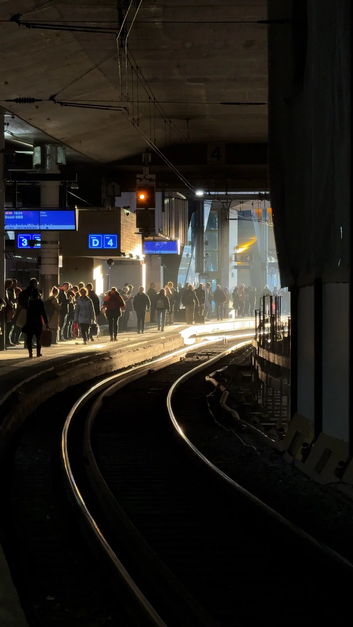 Menschen steigen an einem Bahnsteig in Bern in einen Zug am frühen Morgen ein, mit Gleisen und Signalen, bei Sonnenaufgang.