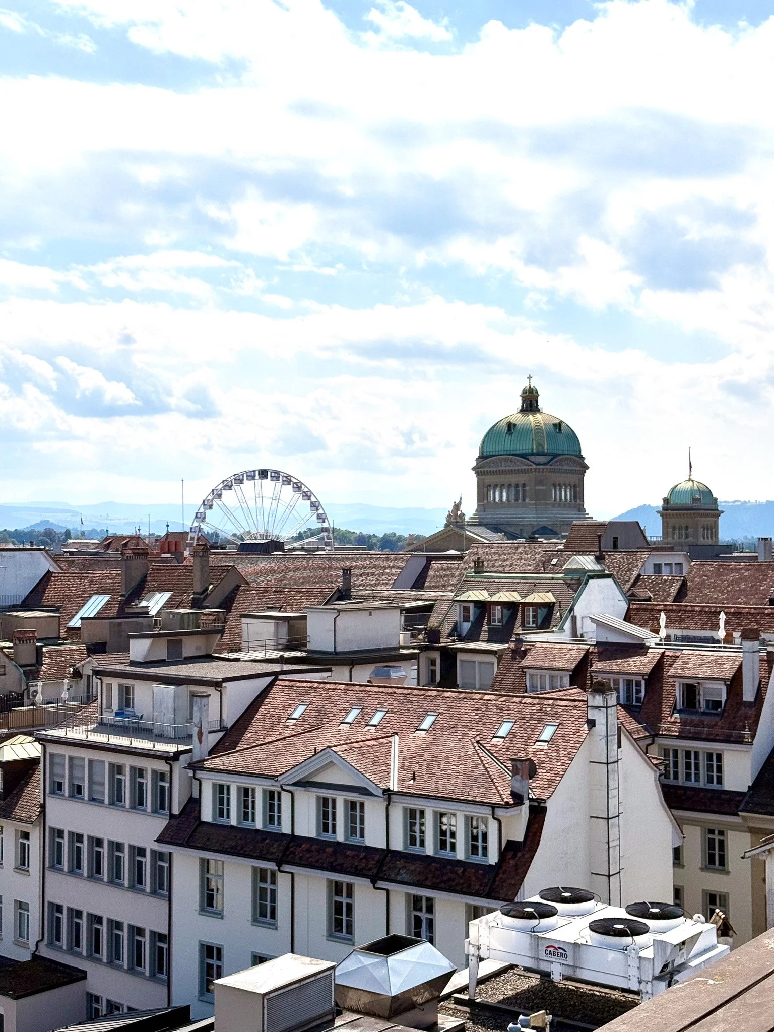 Stadtansicht von Bern mit roten Dächern, Kirchenkuppeln mit grünem Patina-Finish, einem Riesenrad anlässlich der WEURO im Hintergrund und einem blauen Himmel mit Wolken.