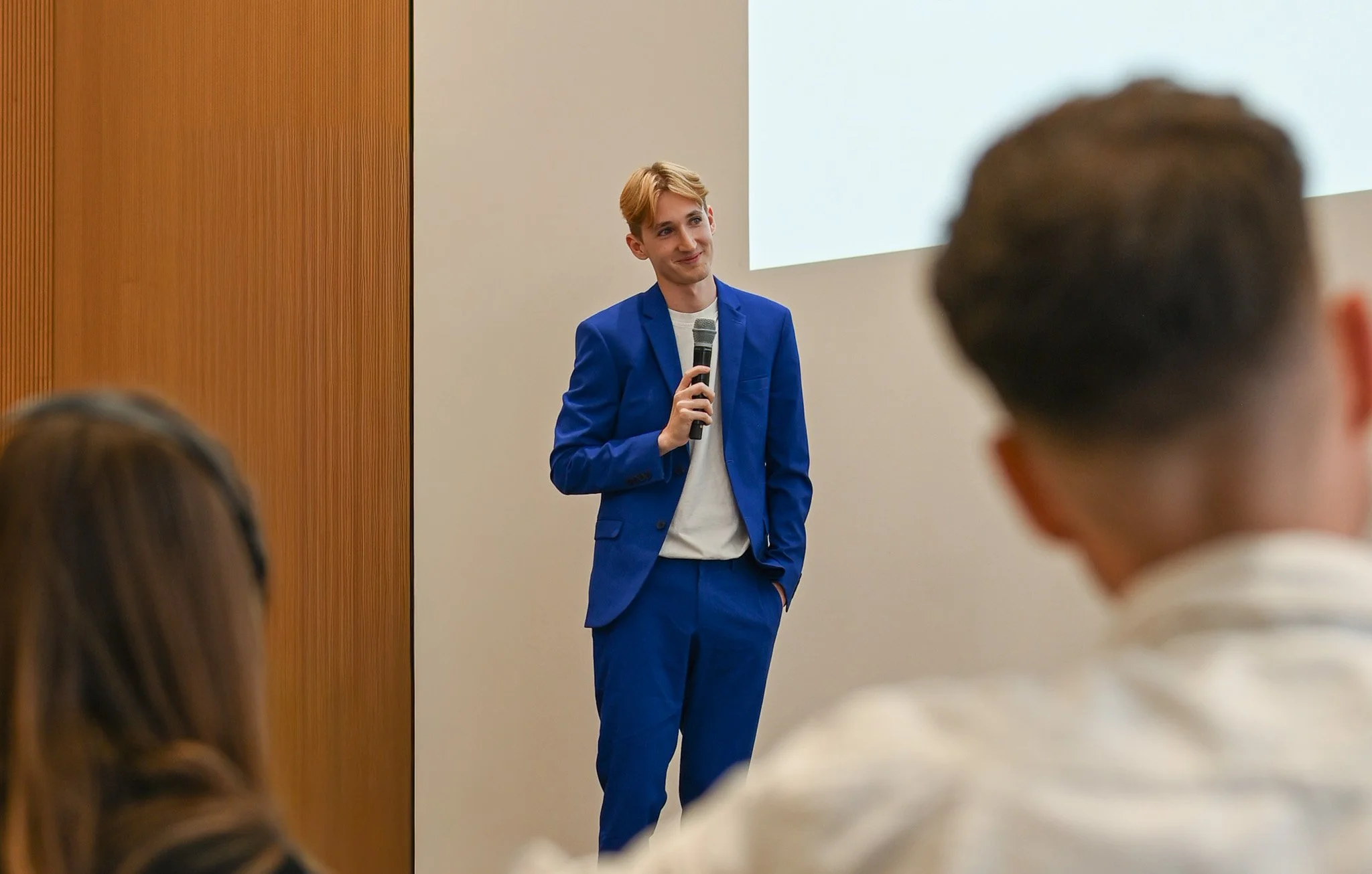 Tim Binda trägt einen blauen Anzug und hält ein Mikrofon, während er vor einer Menschenmasse spricht.