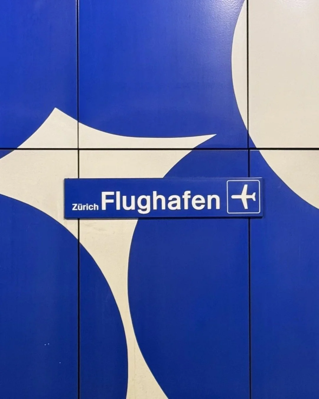 Ein blaues Schild mit weißer Schrift, das auf den Flughafen in Zürich hinweist. Es zeigt die Richtung zum Flughafen mit einem Flugzeug-Symbol.