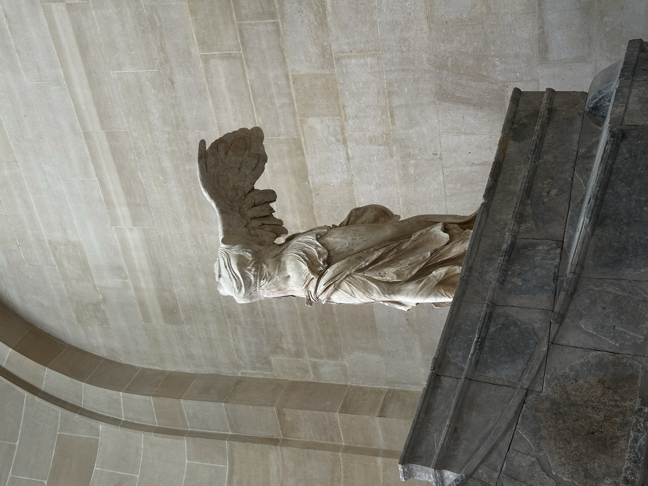 Marble sculpture of a winged female figure missing its head and arms, standing on a stone pedestal against a beige stone wall.
