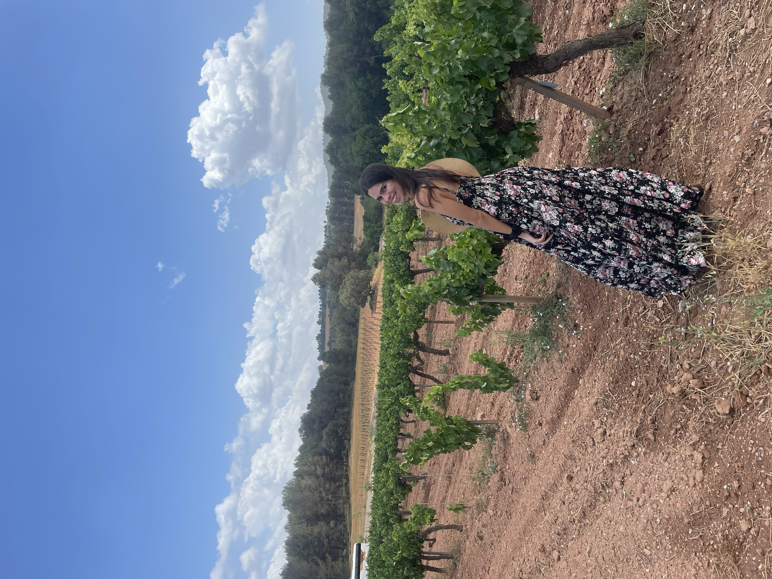 A woman in a black floral dress with a wide-brimmed hat standing in a vineyard on a sunny day with and blue sky and white clouds.