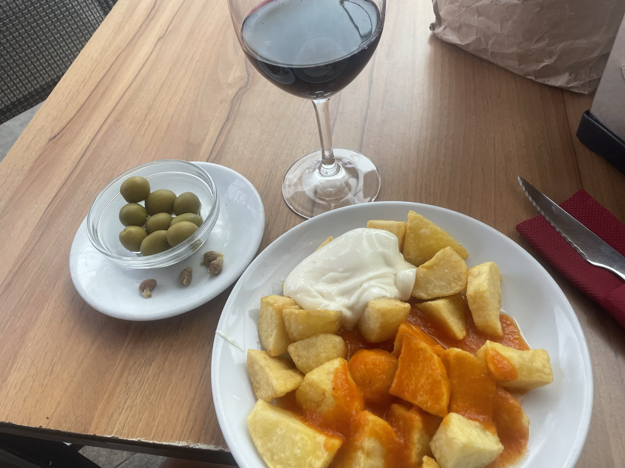 A glass of red wine, a bowl of green olives, and a plate of appetizers including fried potatoes, a fried egg, and some orange vegetables on a wooden table.