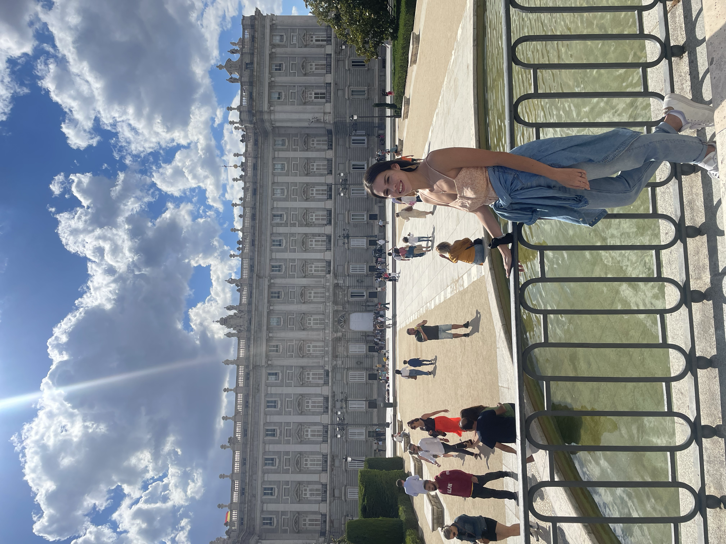 A smiling woman in a pink top, light blue jeans, and white sneakers, standing by a fountain with green water, in front of a grand historic building under a partly cloudy sky.