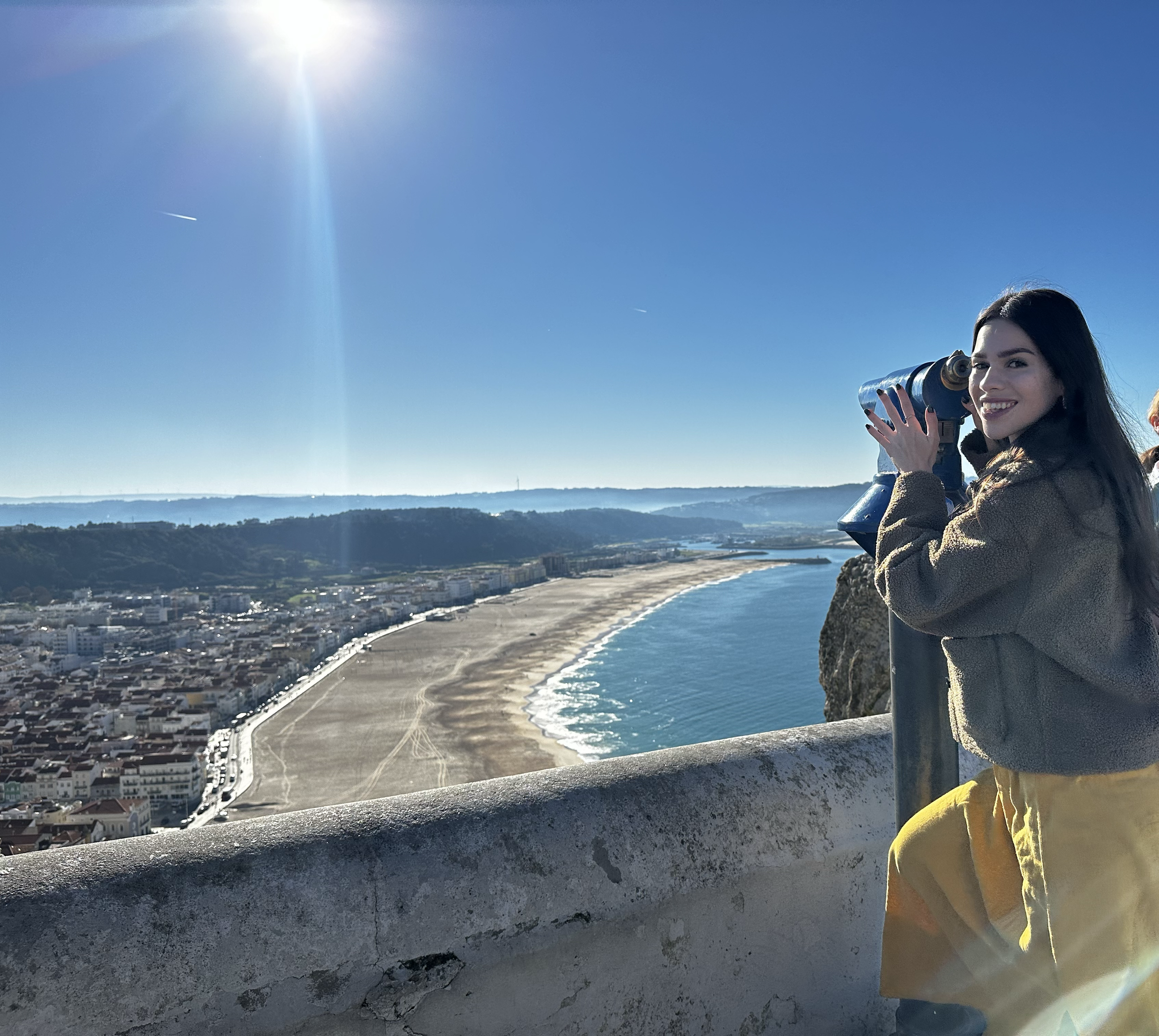 A young woman looking through a telescope at the ocean and coastline on a sunny day.