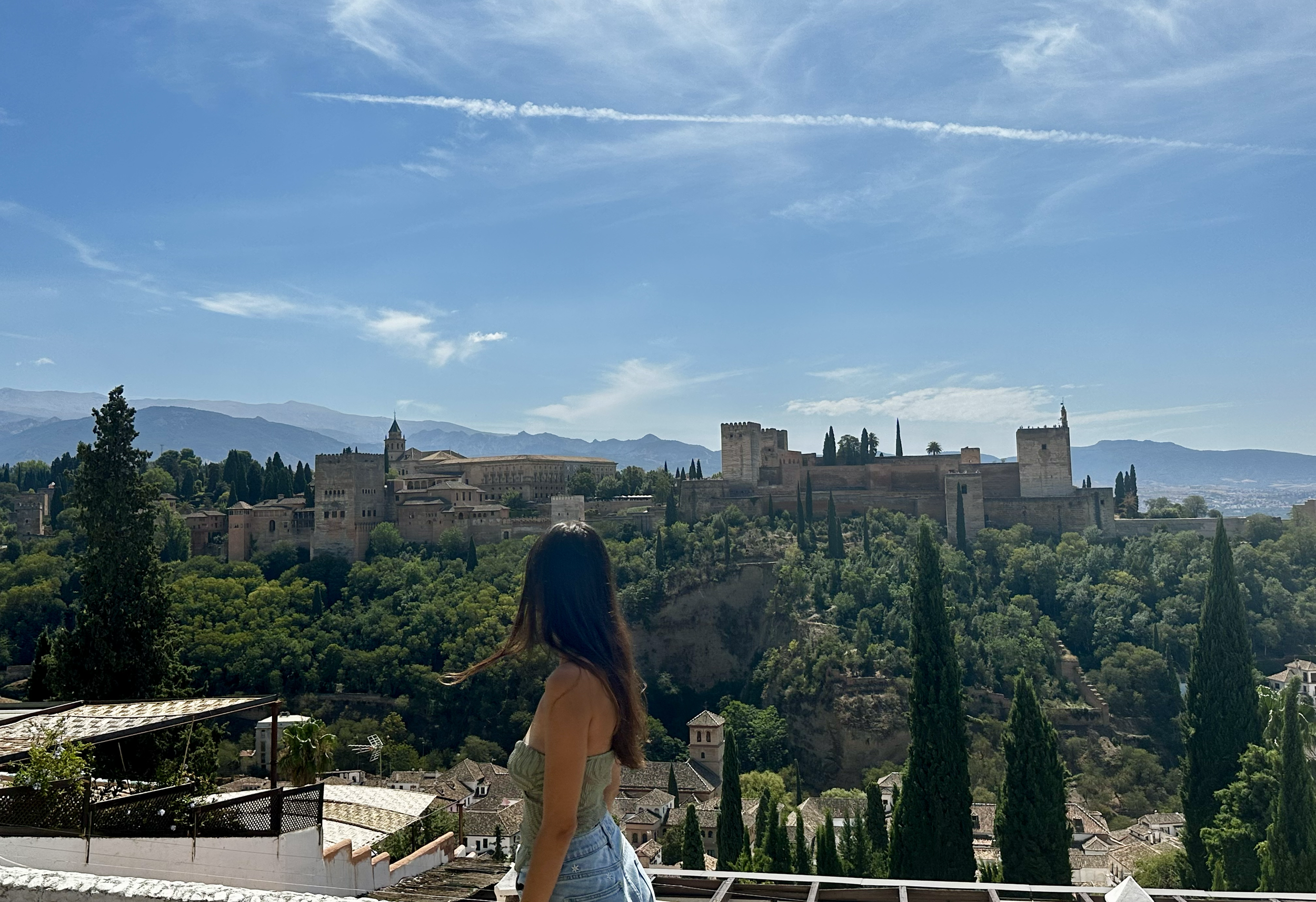 A woman with long hair wearing a beige top and denim shorts standing on a balcony overlooking a scenic view of an ancient city with castles and lush greenery, surrounded by mountains under a blue sky with wispy clouds.