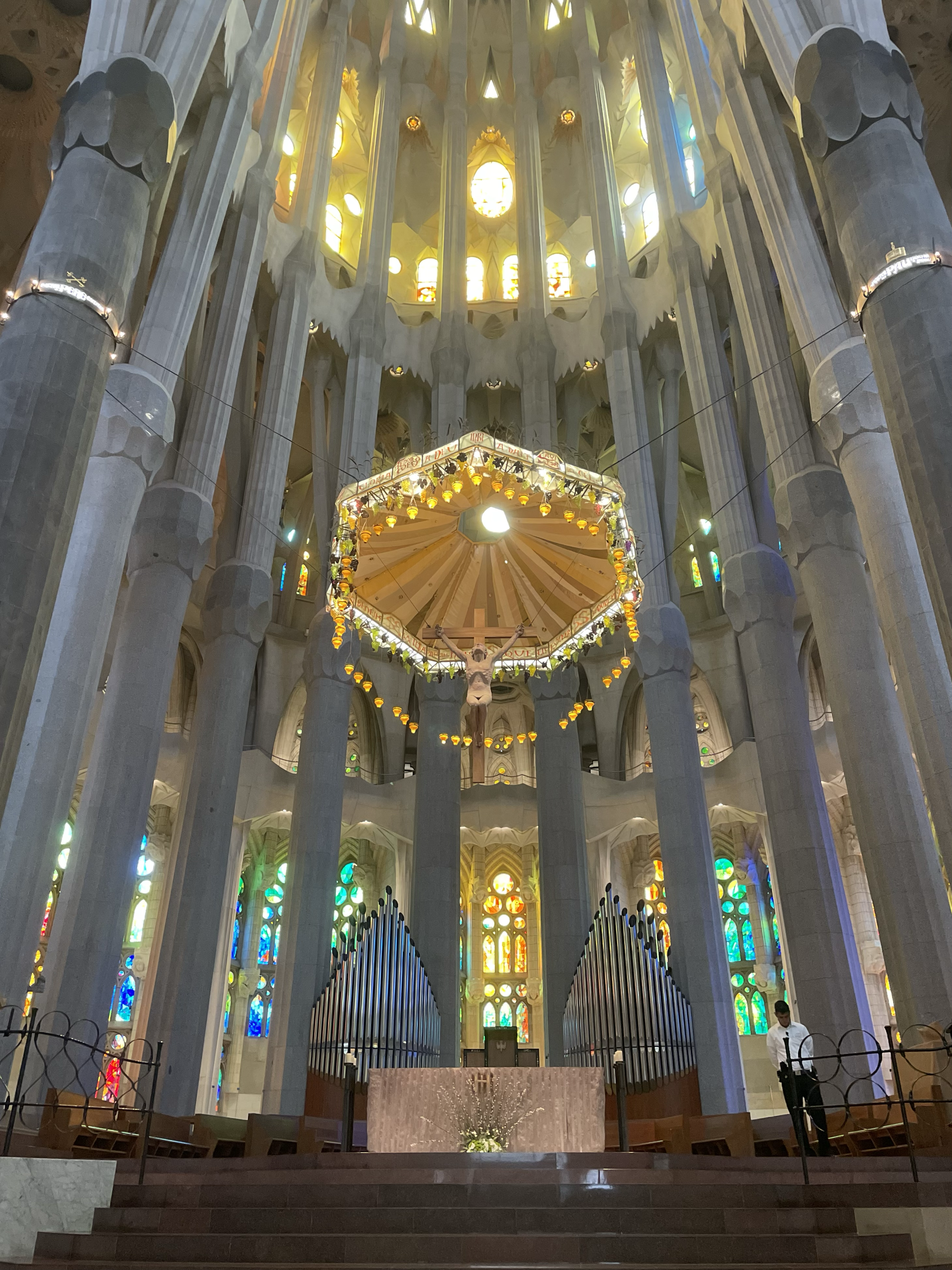 Interior view of Sagrada Família church showing tall columns, colorful stained glass windows, a crucifix with Jesus, and altar with flowers.