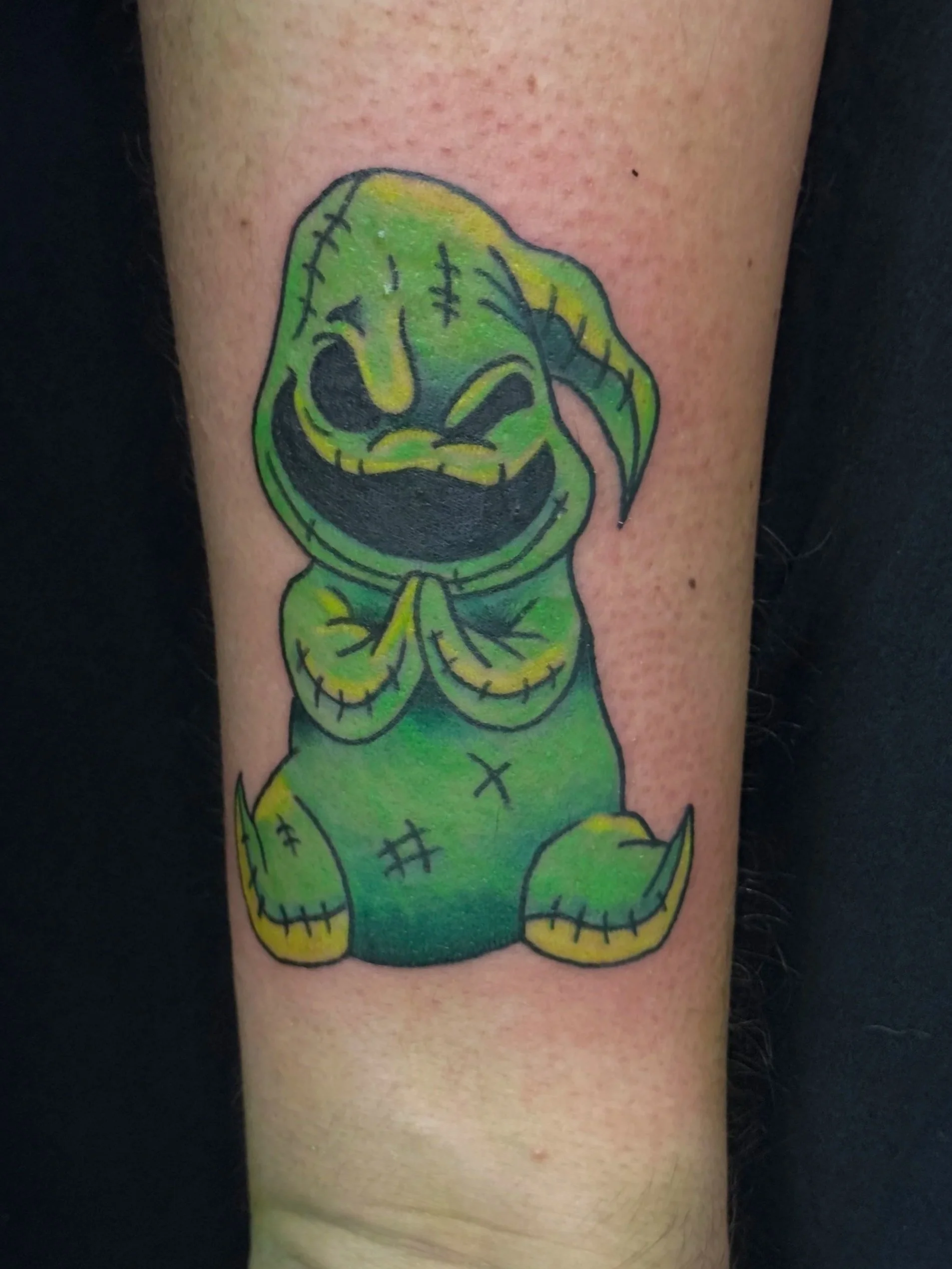 A tattoo of a cartoonish, green, stitched-up zombie creature with a large smile, standing with hands pressed together.