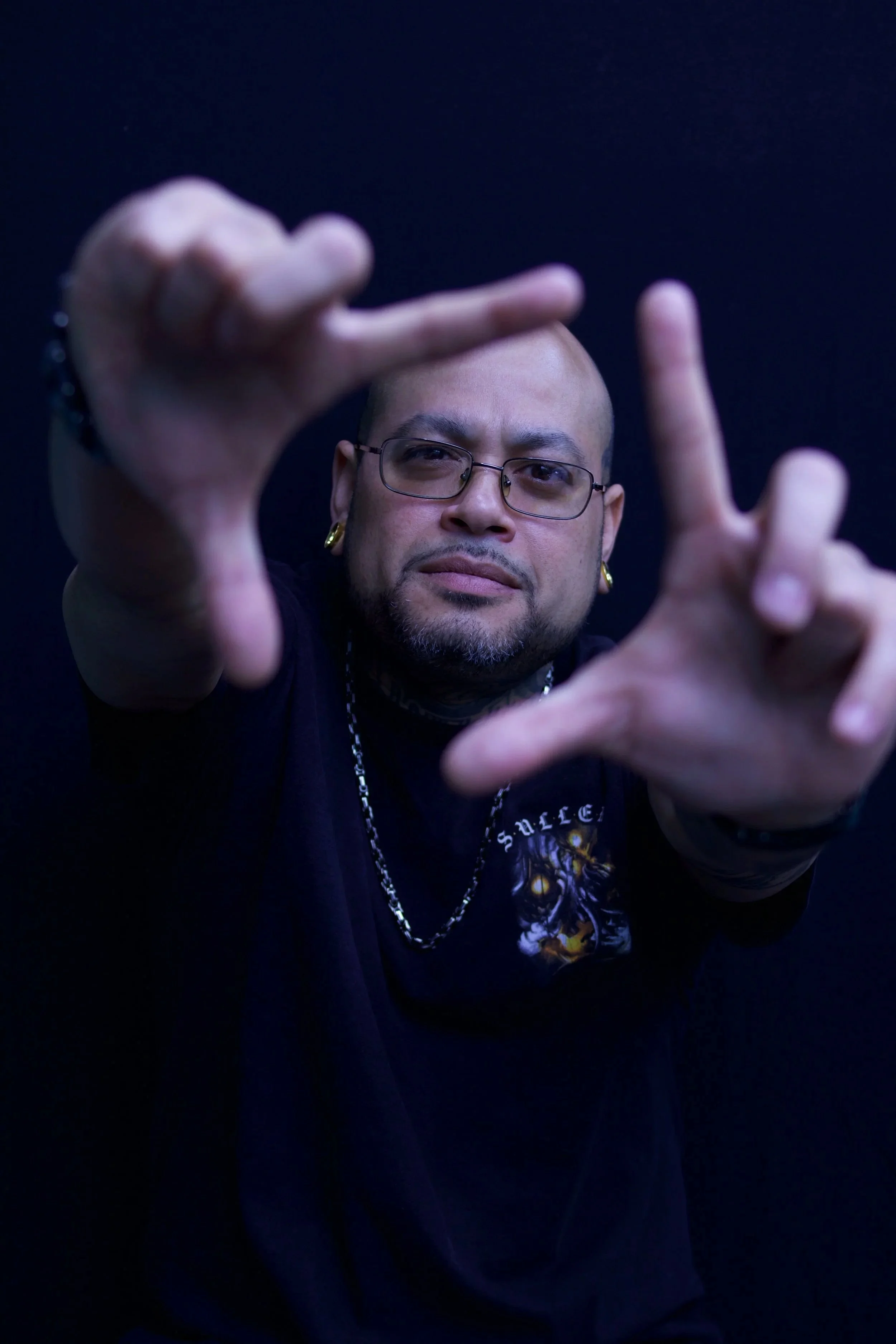 A person with glasses and tribal earrings making a hand gesture against a dark background.