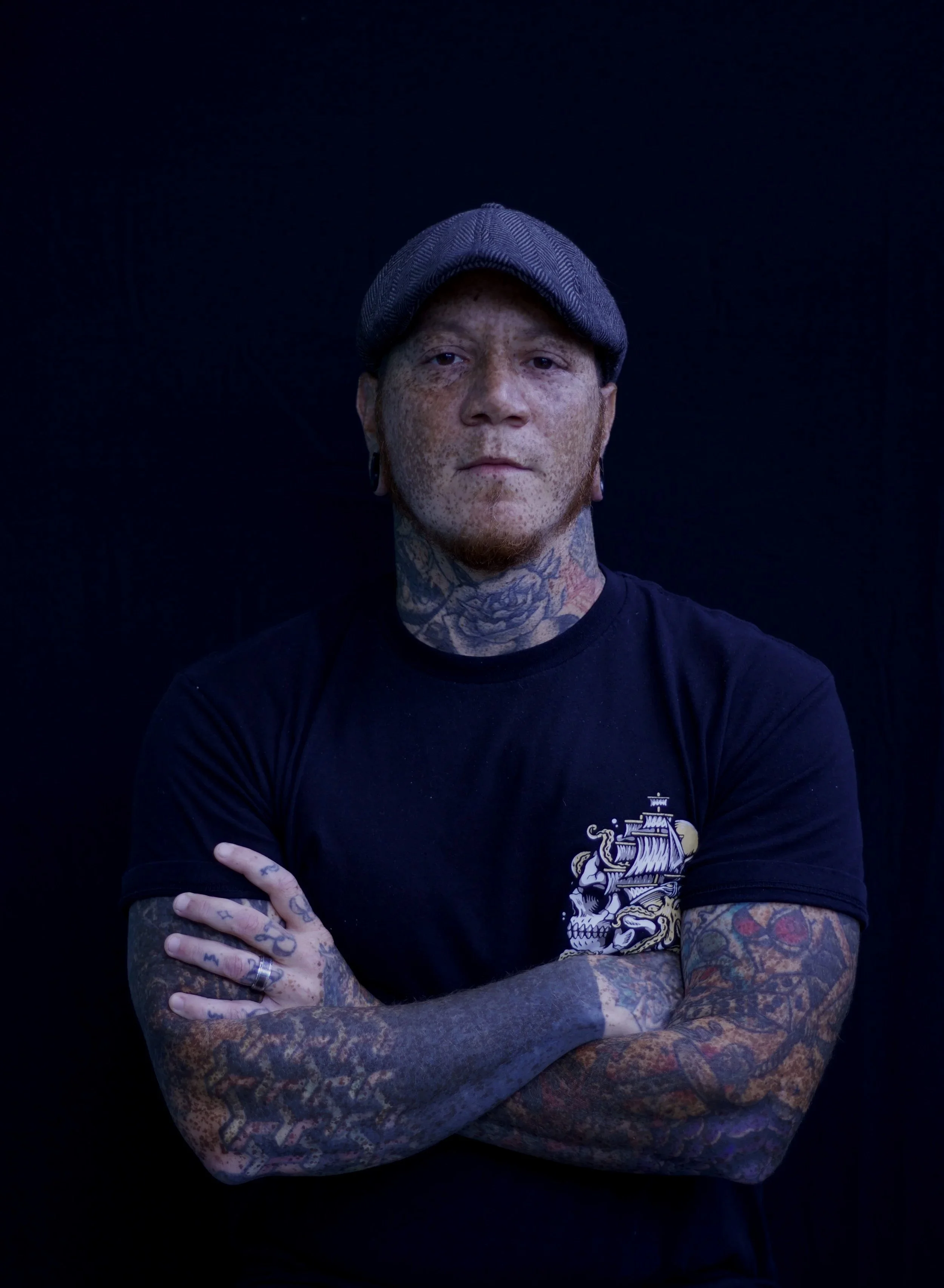 A person with tattoos on their arms and neck, wearing a black t-shirt with a pirate ship graphic, crossed arms, and a gray flat cap, standing against a dark background.