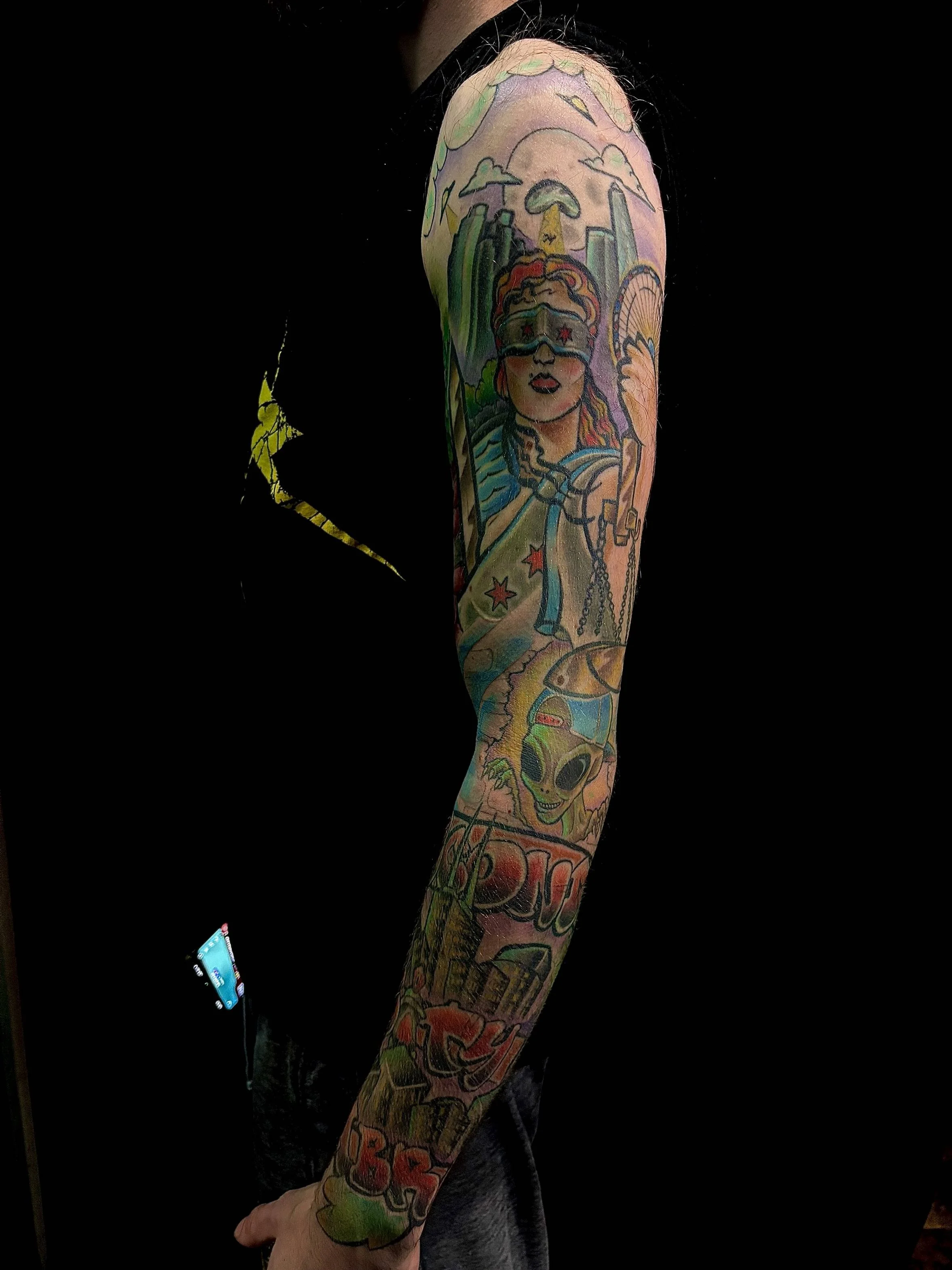 A tattoo sleeve on a person's arm featuring various colorful comic-style images including a woman with sunglasses, a skull, a cityscape, a rainbow, and other abstract designs.