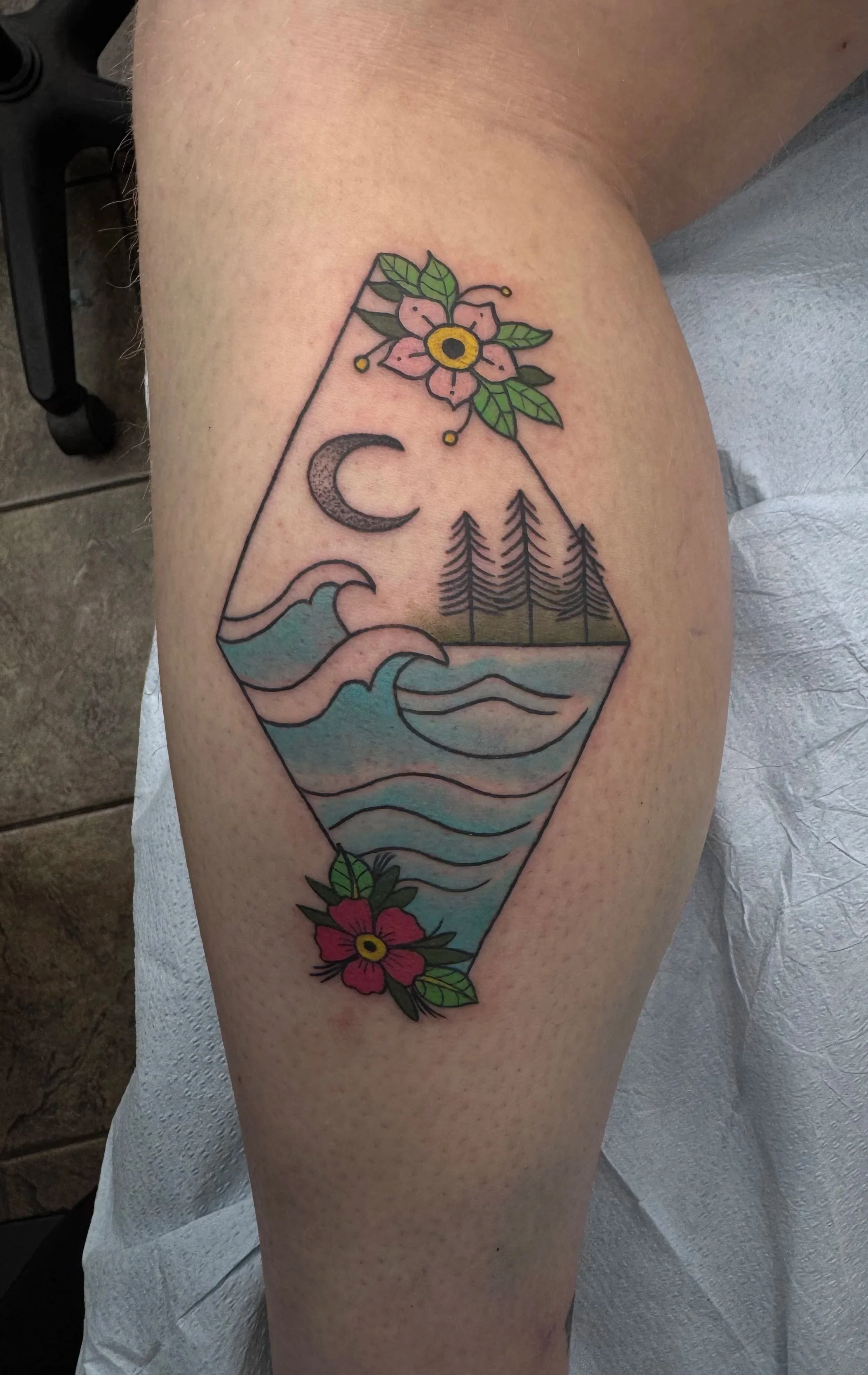 Colorful tattoo on a person's thigh depicting a geometric landscape with mountains, water, pine trees, a crescent moon, and pink and red flowers with green leaves.
