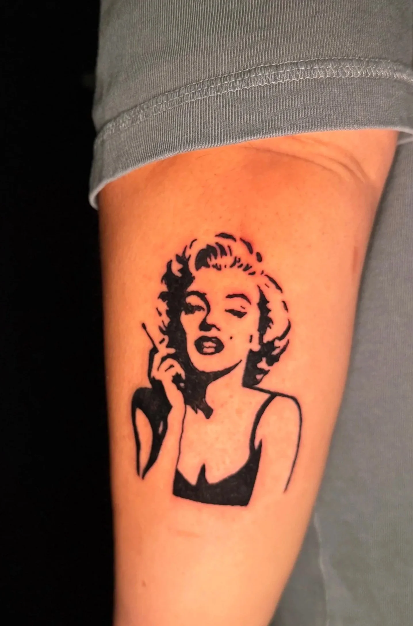 A tattoo of Marilyn Monroe's face on a person's upper arm, with the person's gray sleeve partially visible.