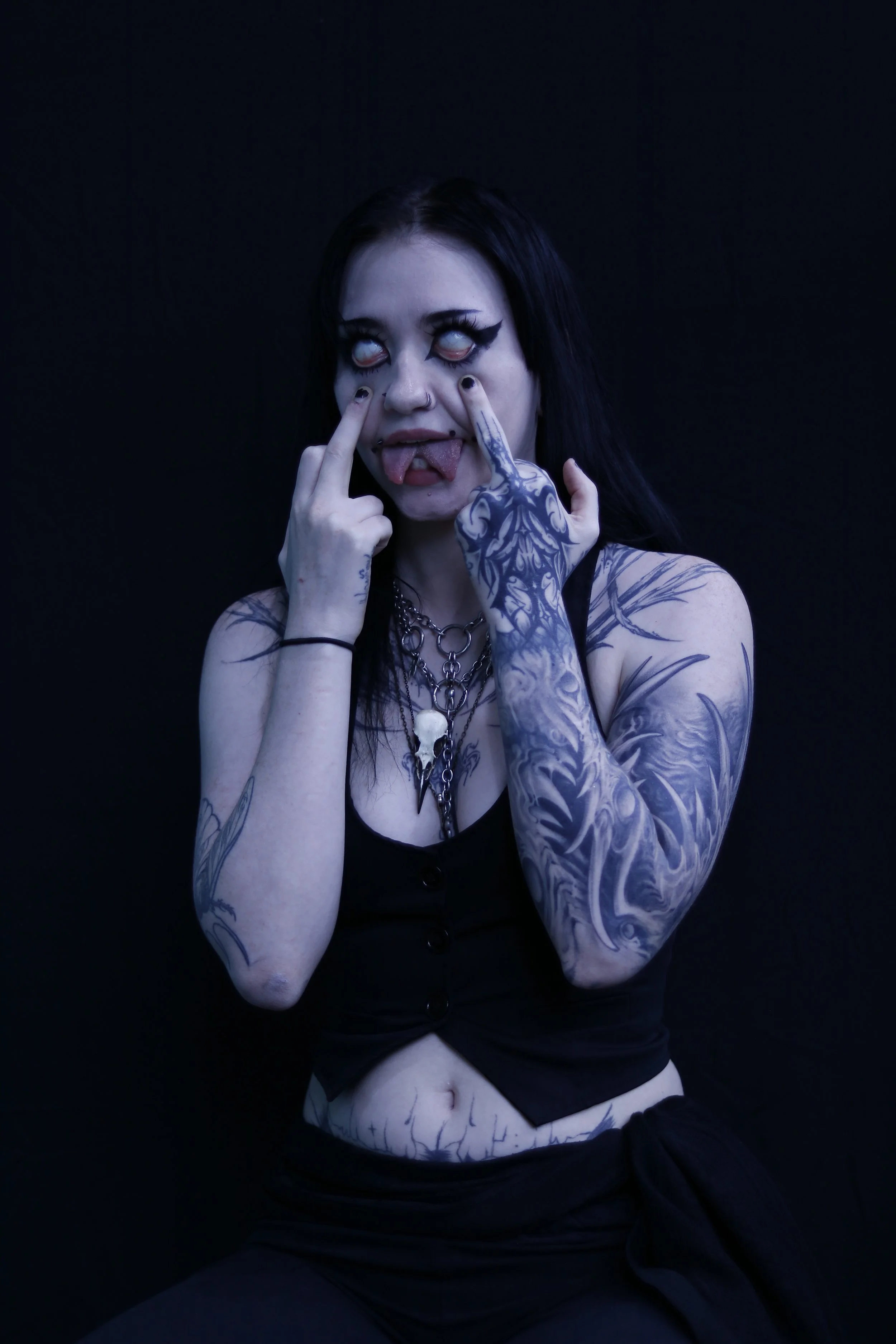 A person with tattoos on their arms and chest, wearing a black top and necklace, making a face with eyes rolled back and tongue out, against a dark background.