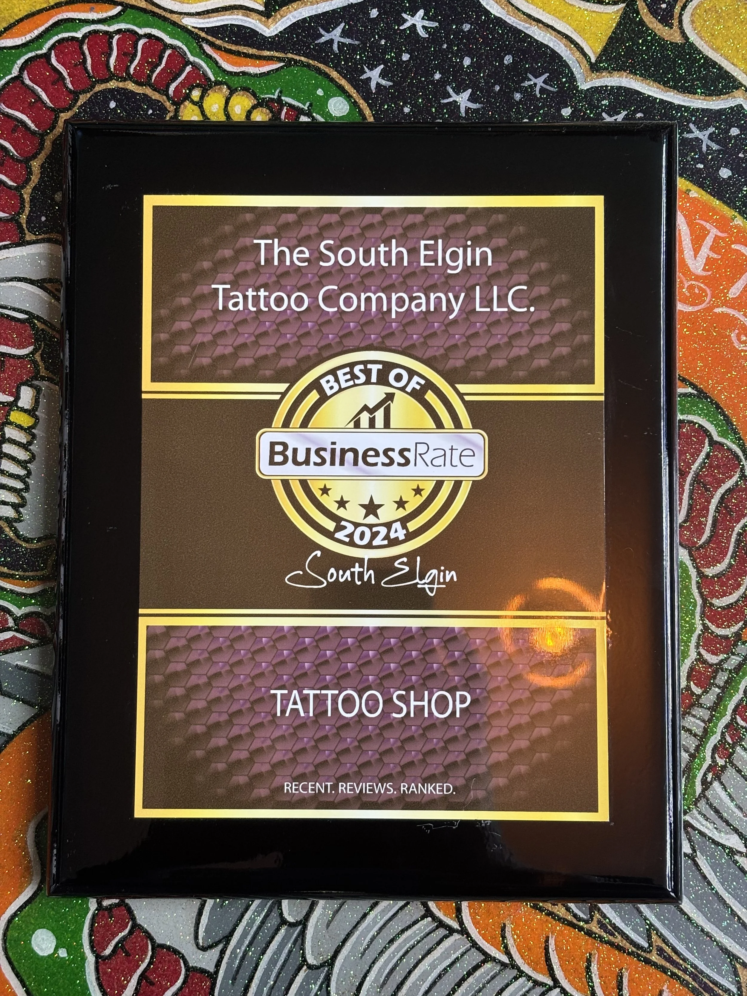 The image shows a framed certificate or award from The South Elgin Tattoo Company LLC, recognizing it as one of the best of 2024. The background features a colorful, glittery, abstract pattern with red, green, orange, yellow, and black colors.