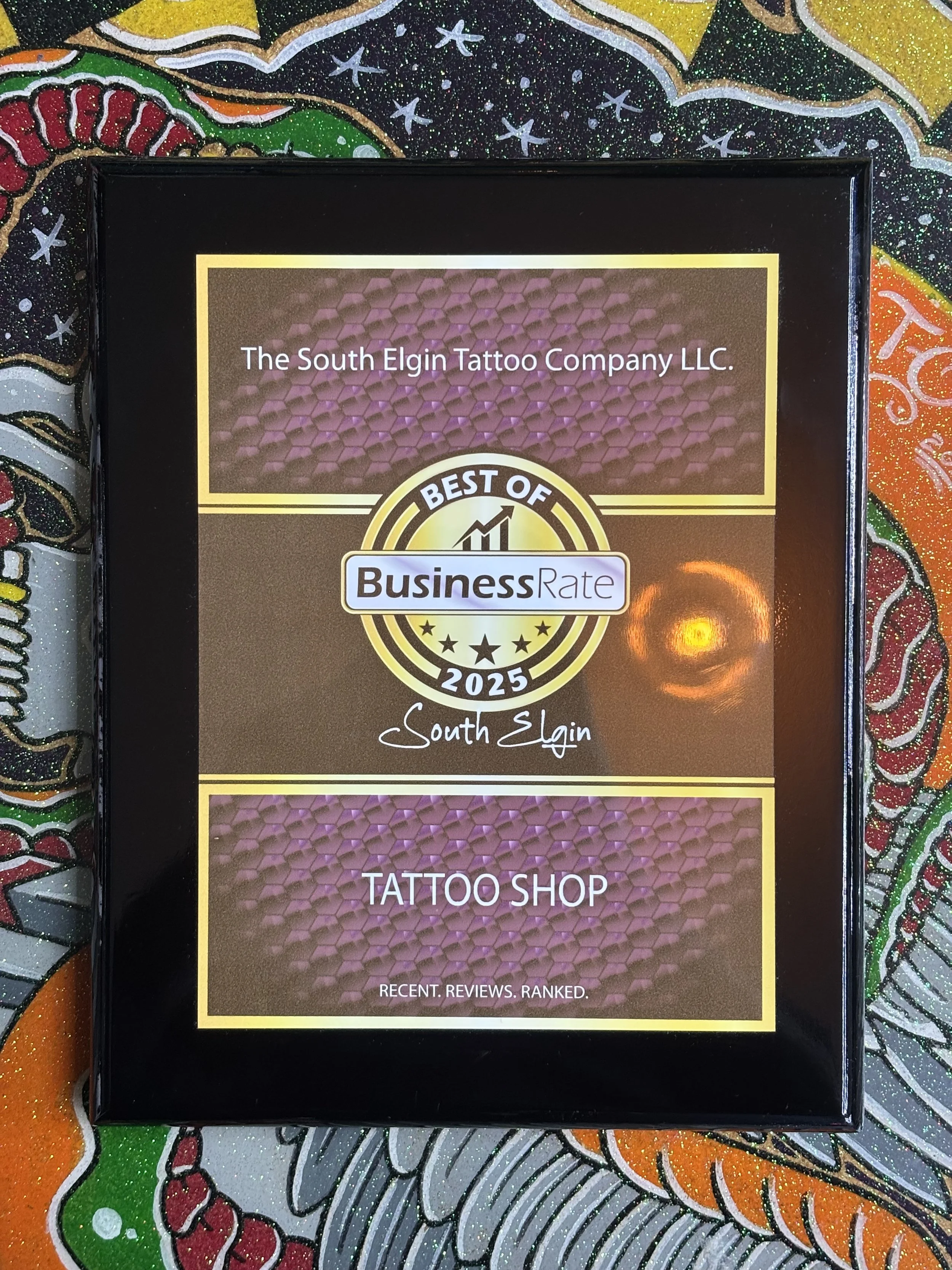 Close-up of a tattoo shop's award or recognition plaque with the text 'The South Elgin Tattoo Company LLC,' 'Best of Business Rate 2025,' and 'South Elgin.' The plaque has a border with a honeycomb pattern and is placed on a colorful, glittery background with floral and abstract patterns.