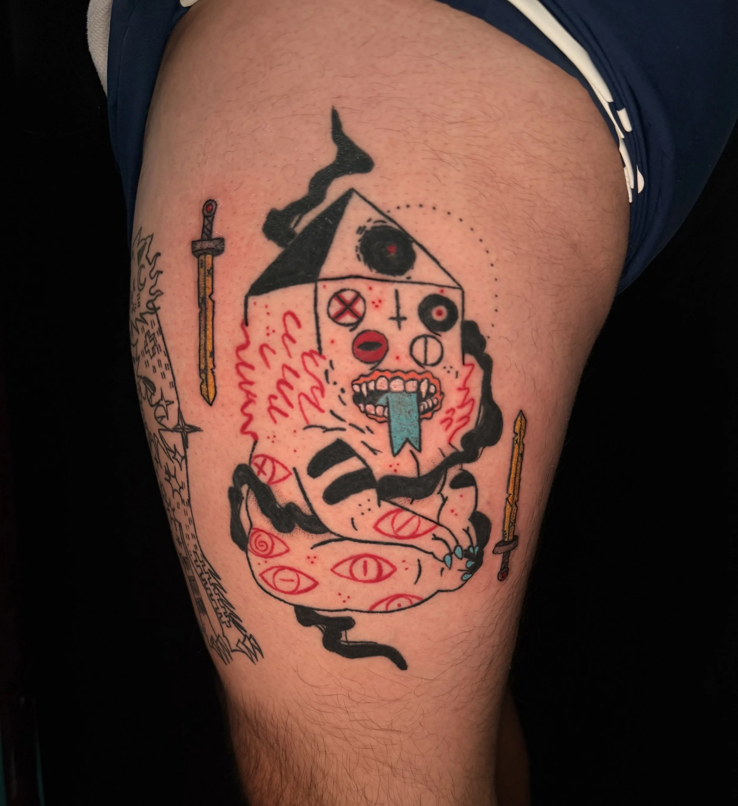 Tattoo of a stylized skull with tribal elements on person's upper arm, with a blue tongue-like extension and red accent details, surrounded by other tattoos and a person's skin.
