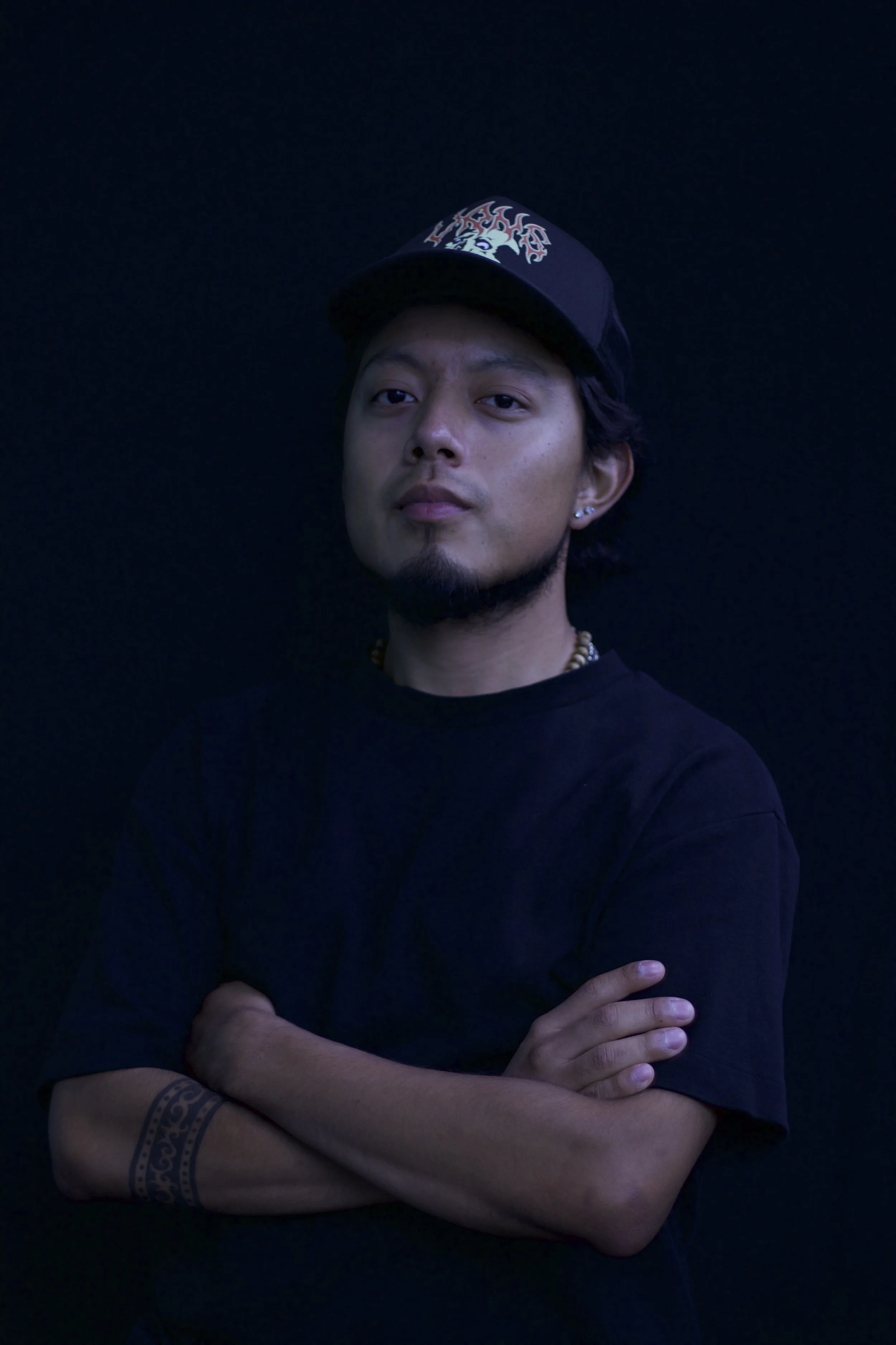 A man with medium-length dark hair, a goatee, and earrings, wearing a black T-shirt and a black baseball cap with a graphic design, standing against a black background with arms crossed.