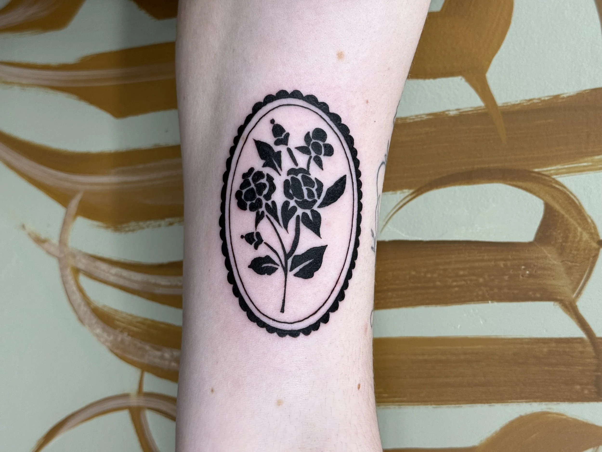 Tattoo of a black outline of a flowering plant inside an oval border on a person's forearm. The background is a green wall with a wood pattern.