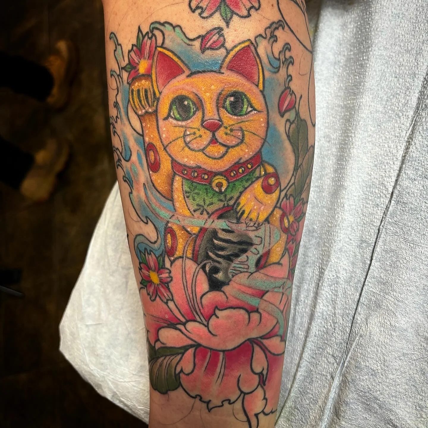 A colorful tattoo of a lucky cat with big green eyes, wearing a collar with a bell, surrounded by pink and red flowers, on a person's arm.
