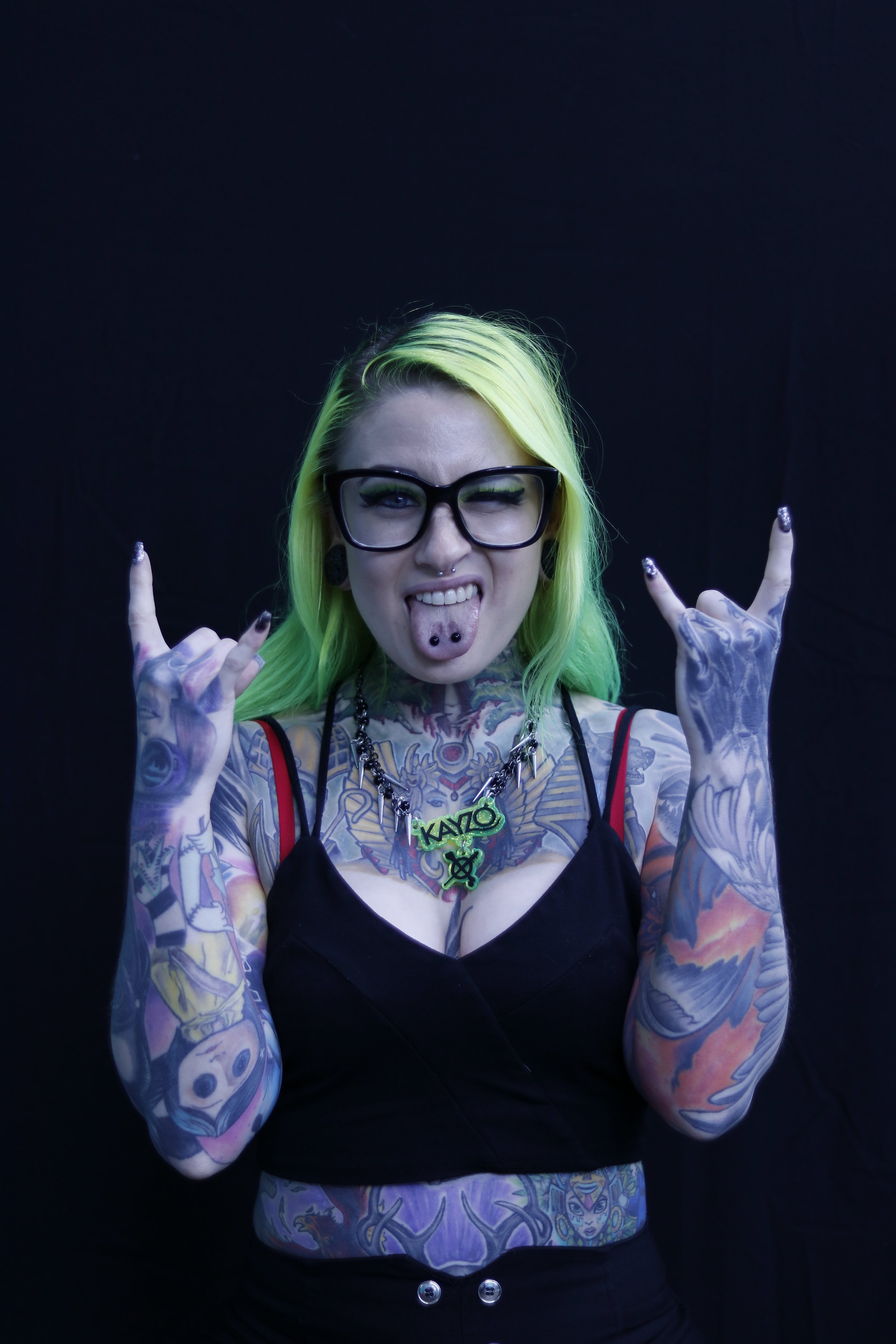 A young woman with neon green hair and glasses making a rock and roll gesture. She is sticking out her tongue with a tongue piercing. She has tattoos on her arms and chest, and is wearing a necklace with a green pendant. She is dressed in a black top, and the background is plain black.