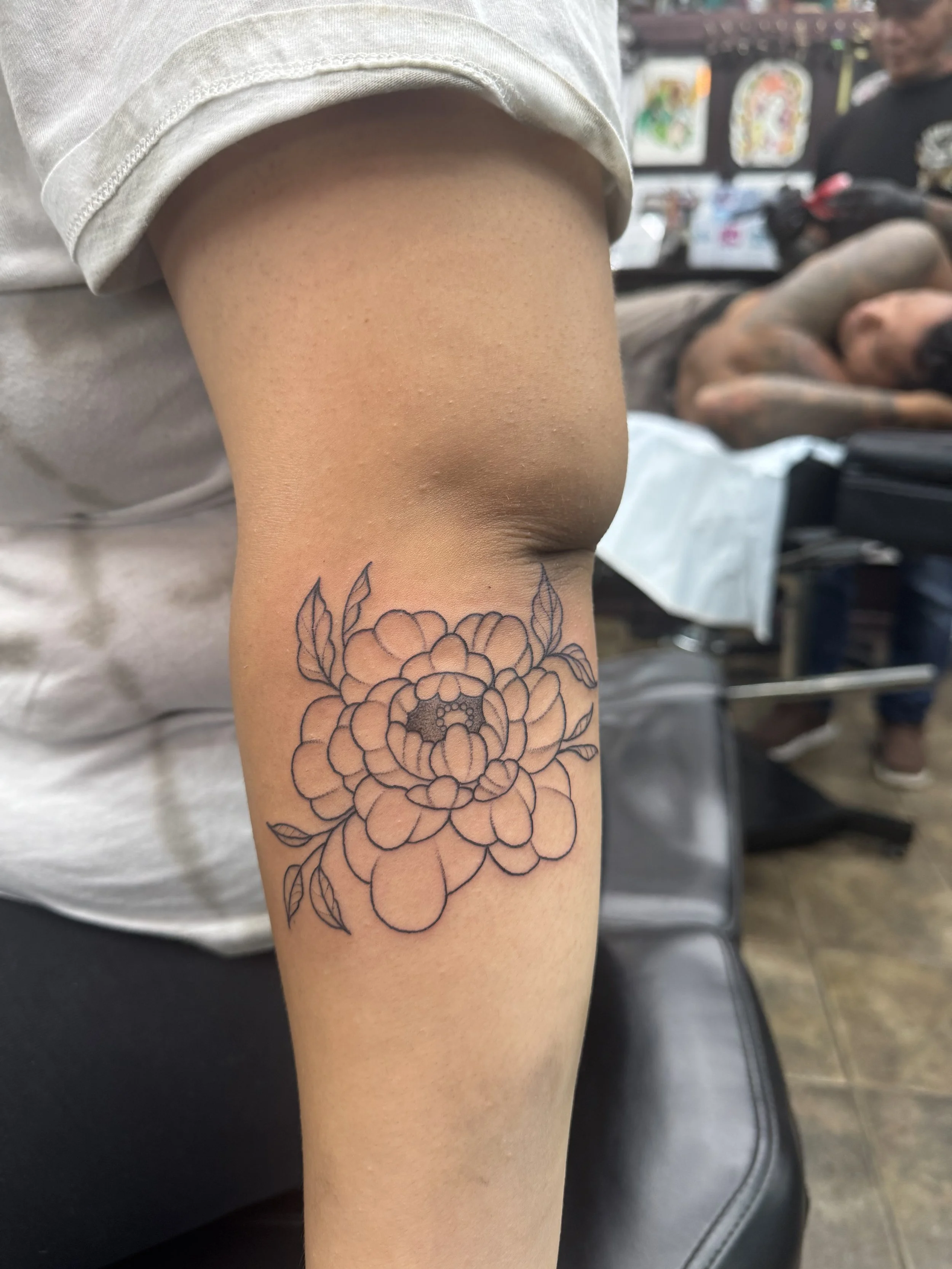 A person's arm with a floral tattoo, wearing a short sleeve shirt, in a tattoo or art shop with artworks on the wall and people in the background.