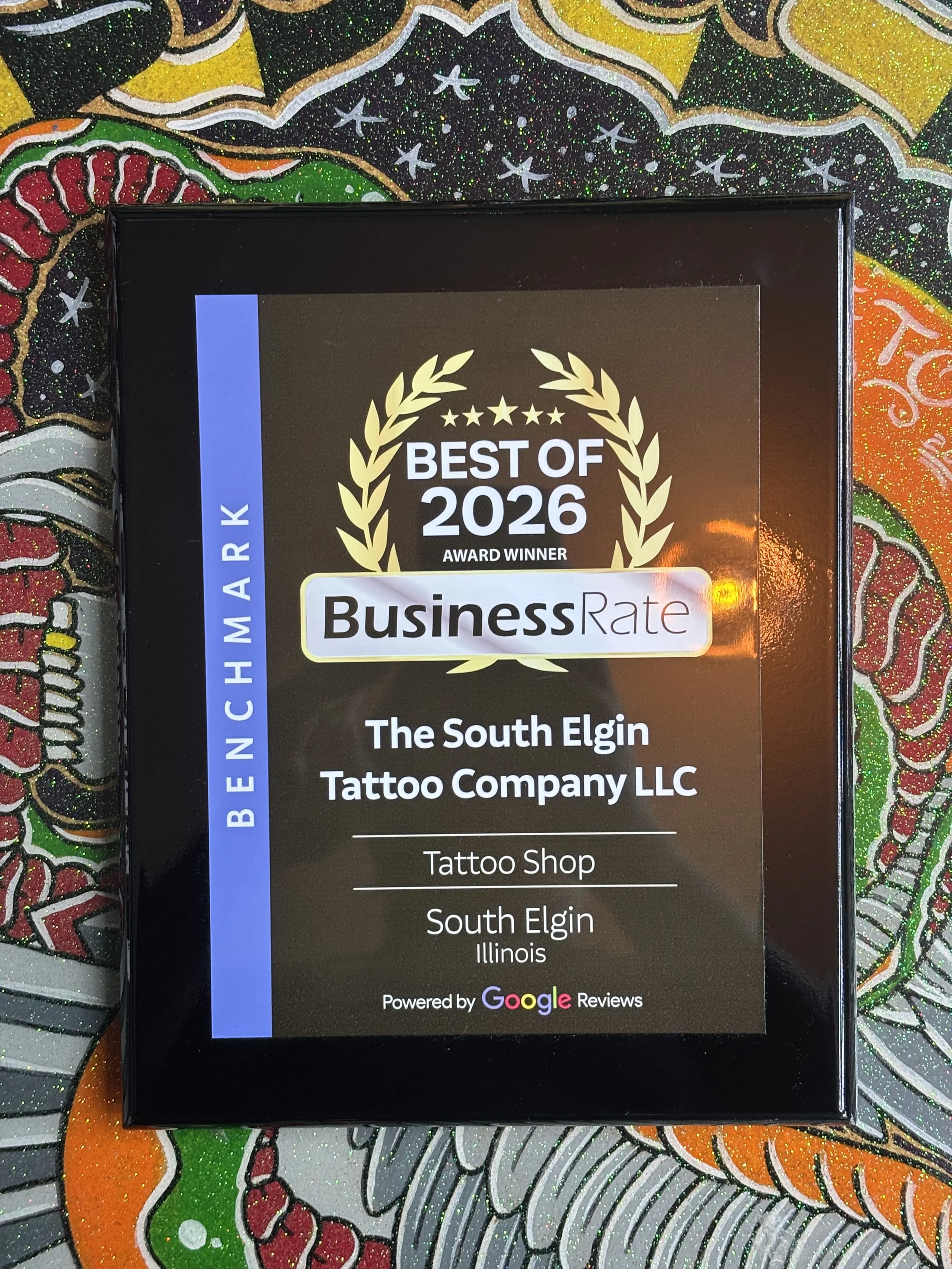 Award plaque with the text "Best of 2026, Award Winner, Business Rate," presented to The South Elgin Tattoo Company LLC in Illinois, with a decorative gold laurel wreath and star design, placed on a colorful, glittery, and patterned background.