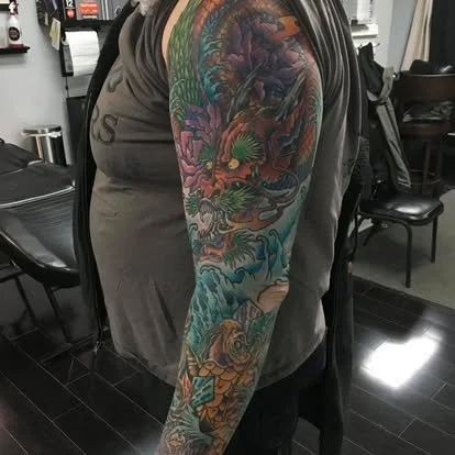 A person's arm with a colorful full sleeve tattoo featuring dragons, waves, and mythical creatures in vivid shades of green, purple, and orange.