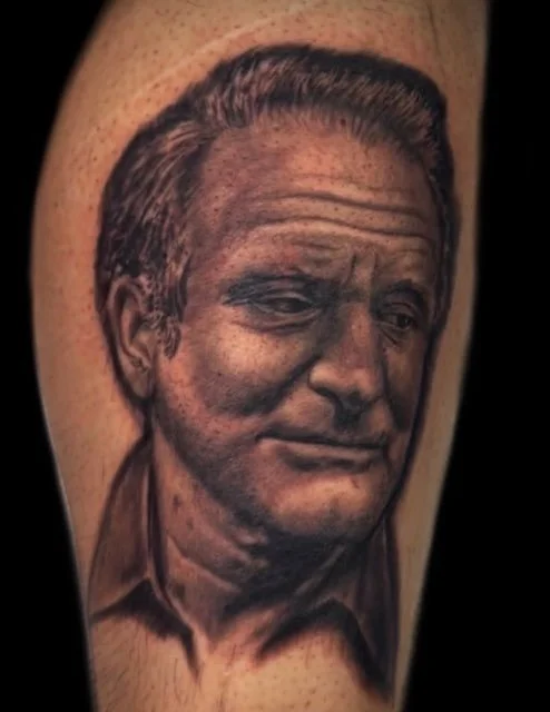Realistic black and gray tattoo of a man's face with a slight smile, depicting detailed facial features and hair, on a person's arm.