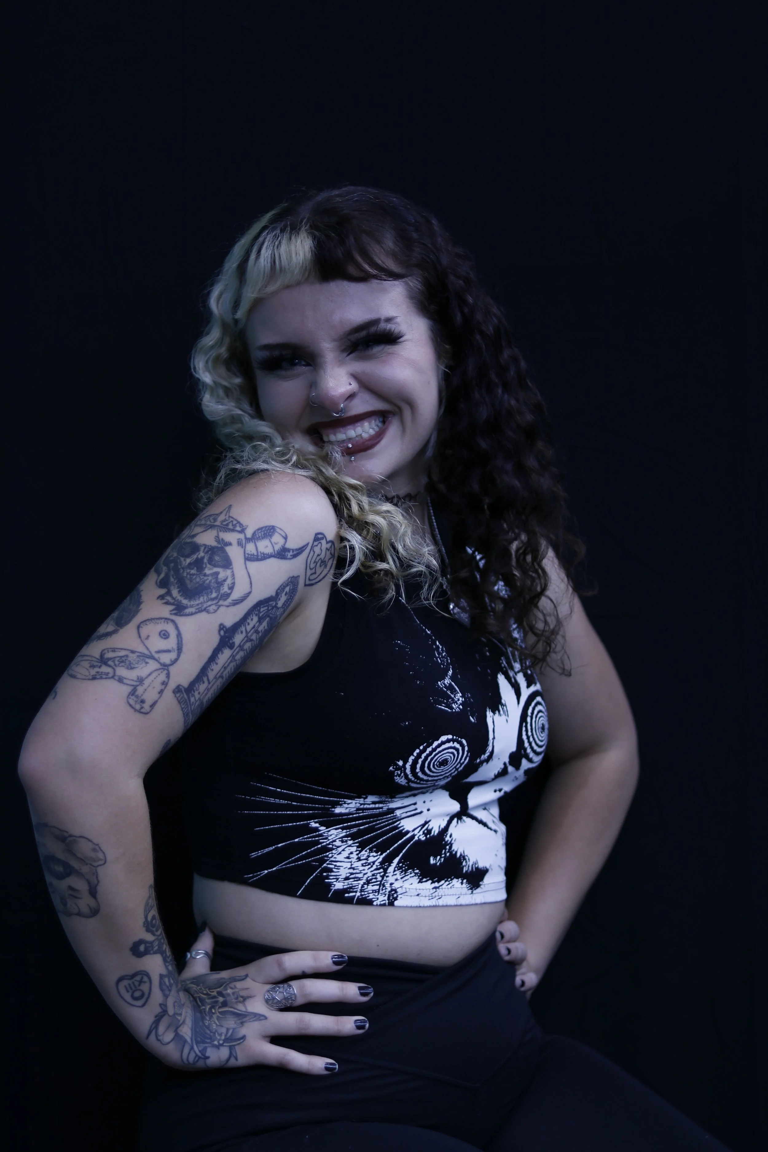 A young woman with dark curly hair and blonde streaks, smiling and showing her tattoos on her arm and side, wearing a black and white graphic crop top, standing against a dark background.