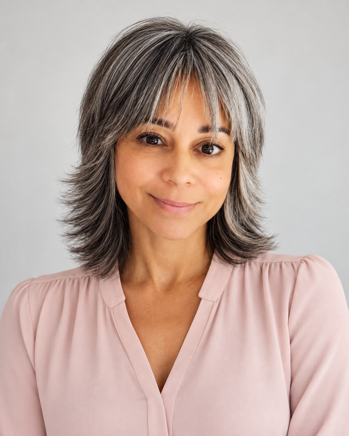 Portrait of a middle-aged woman with short, gray hair, smiling, wearing a light pink blouse, standing against a plain light background.