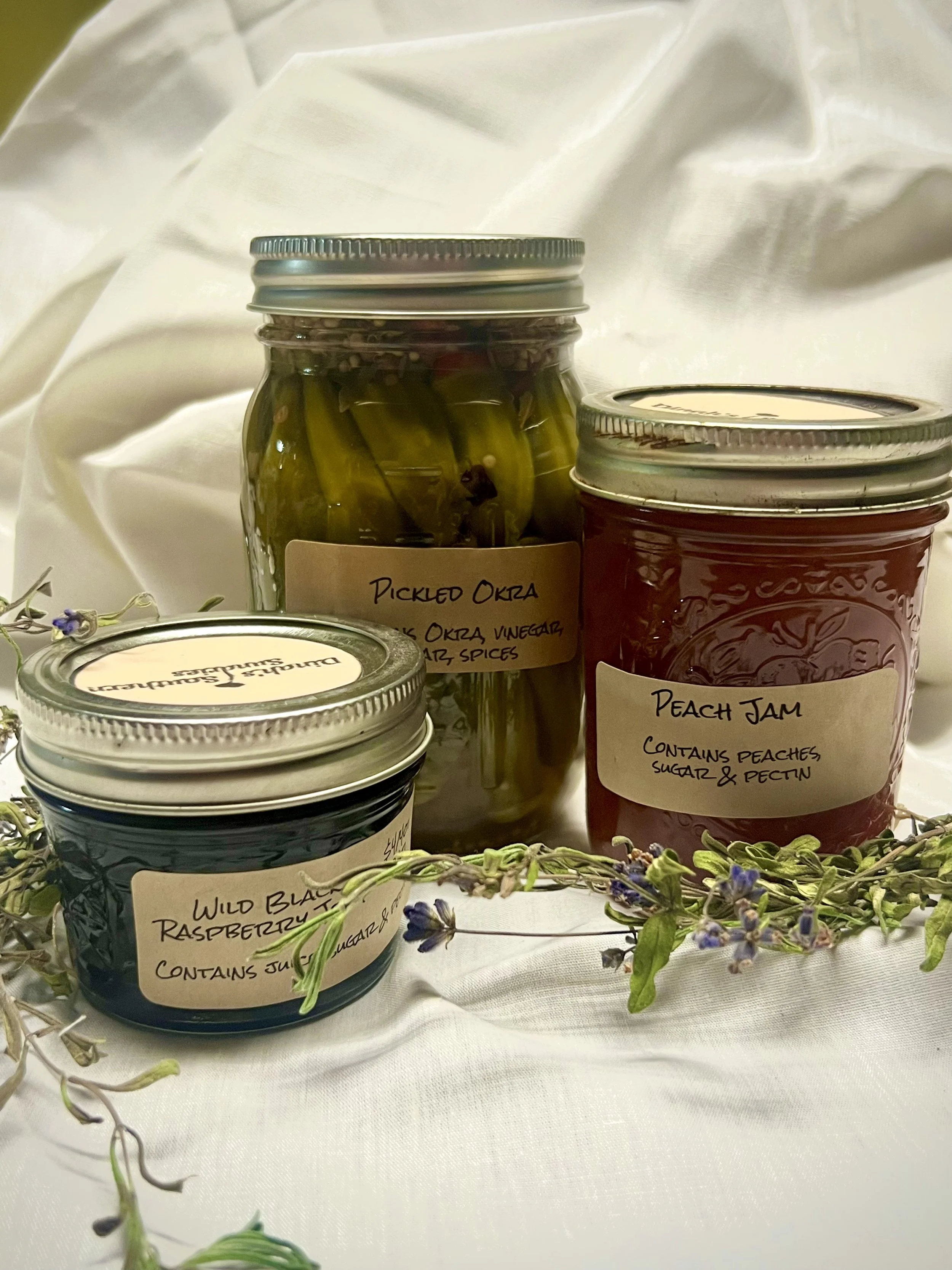Hand-labeled jars of pickled okra, peach jam, and wild black raspberry preserves arranged on a white cloth with flowers.