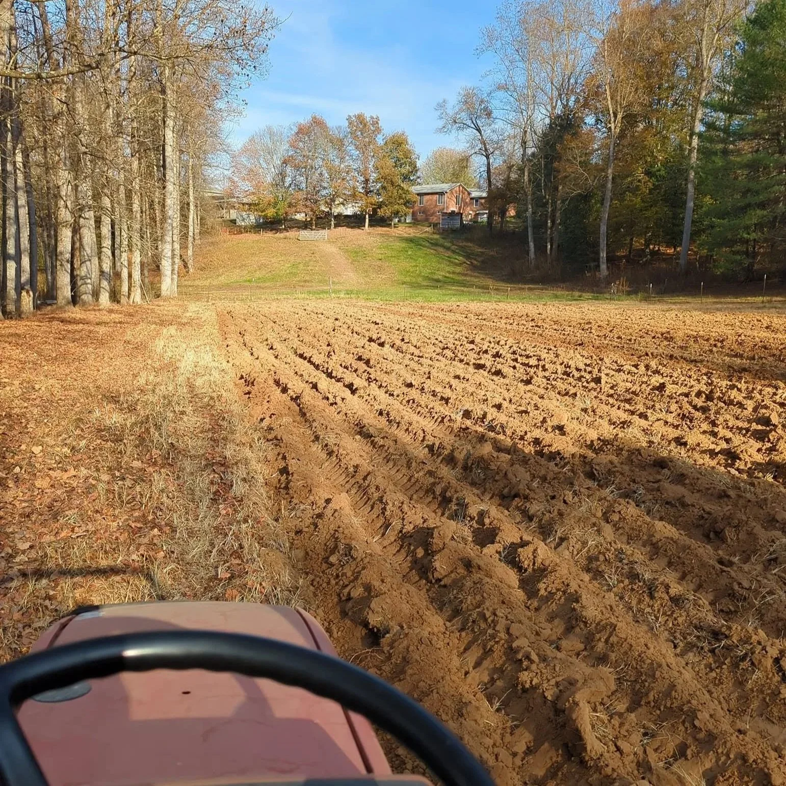 View from a tractor across a freshly plowed field toward a wooded rural home in autumn