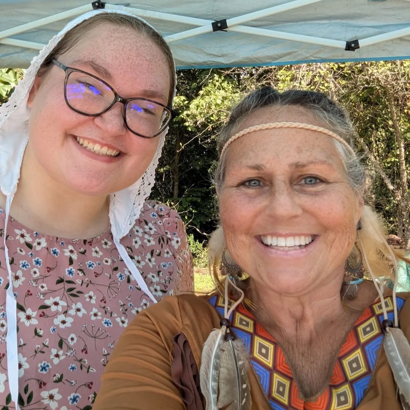 Two smiling women pose for a selfie outdoors under a canopy, wearing historical-style clothing and accessories.