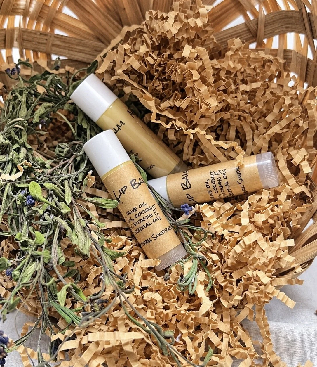 Handmade herbal lip balm tubes surrounded by dried herbs and crinkle paper packaging